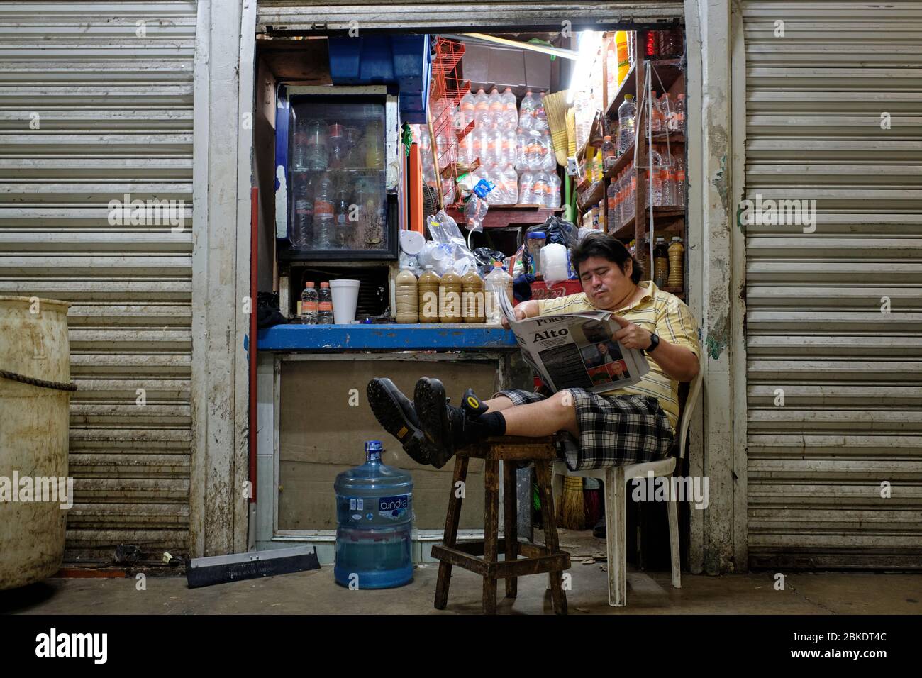 Uomo che legge il giornale seduto con le gambe distese su uno sgabello di fronte ad un piccolo negozio nel mercato comunale di Merida. Foto Stock