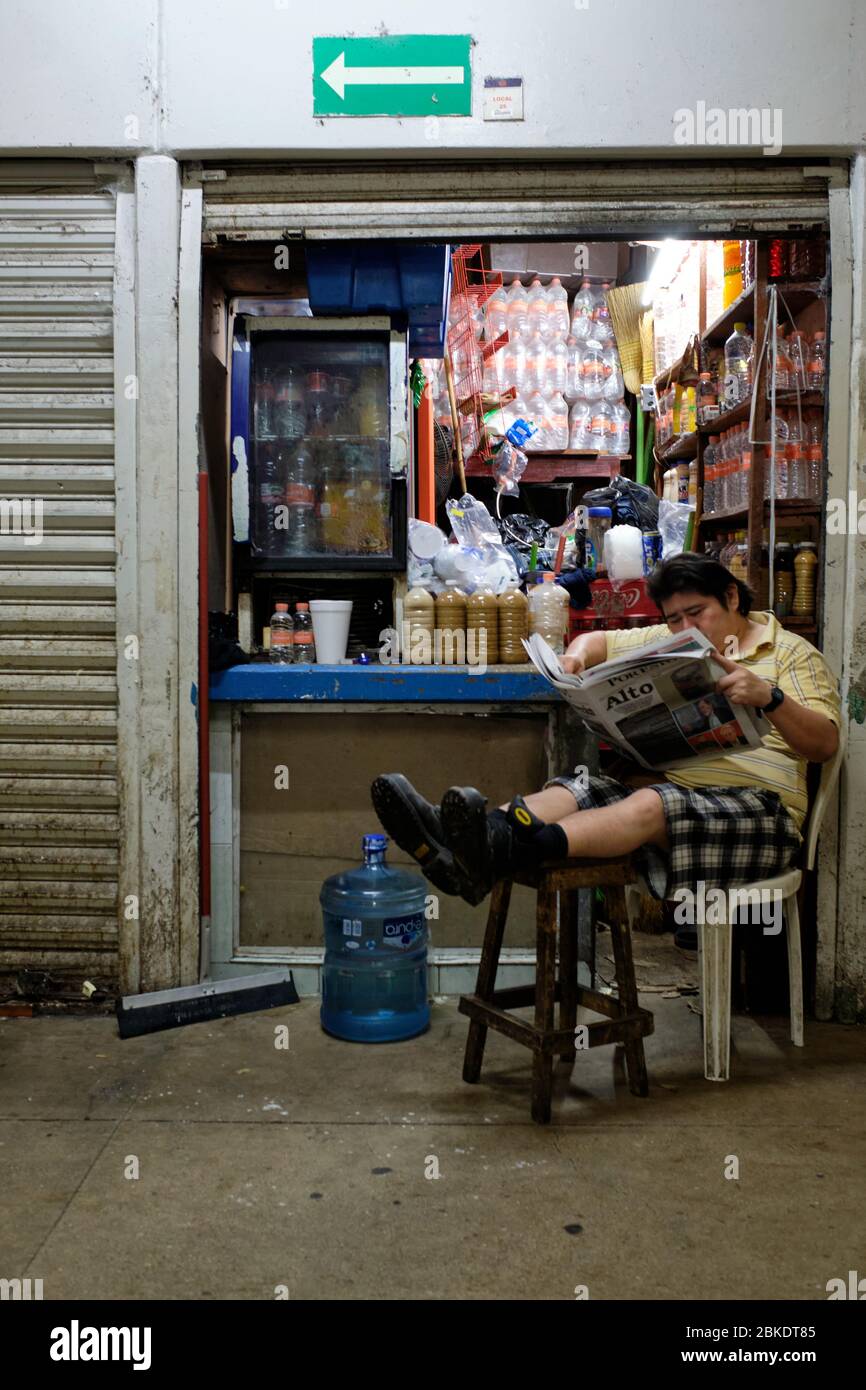 Uomo che legge il giornale seduto con le gambe distese su uno sgabello di fronte ad un piccolo negozio nel mercato comunale di Merida. Foto Stock