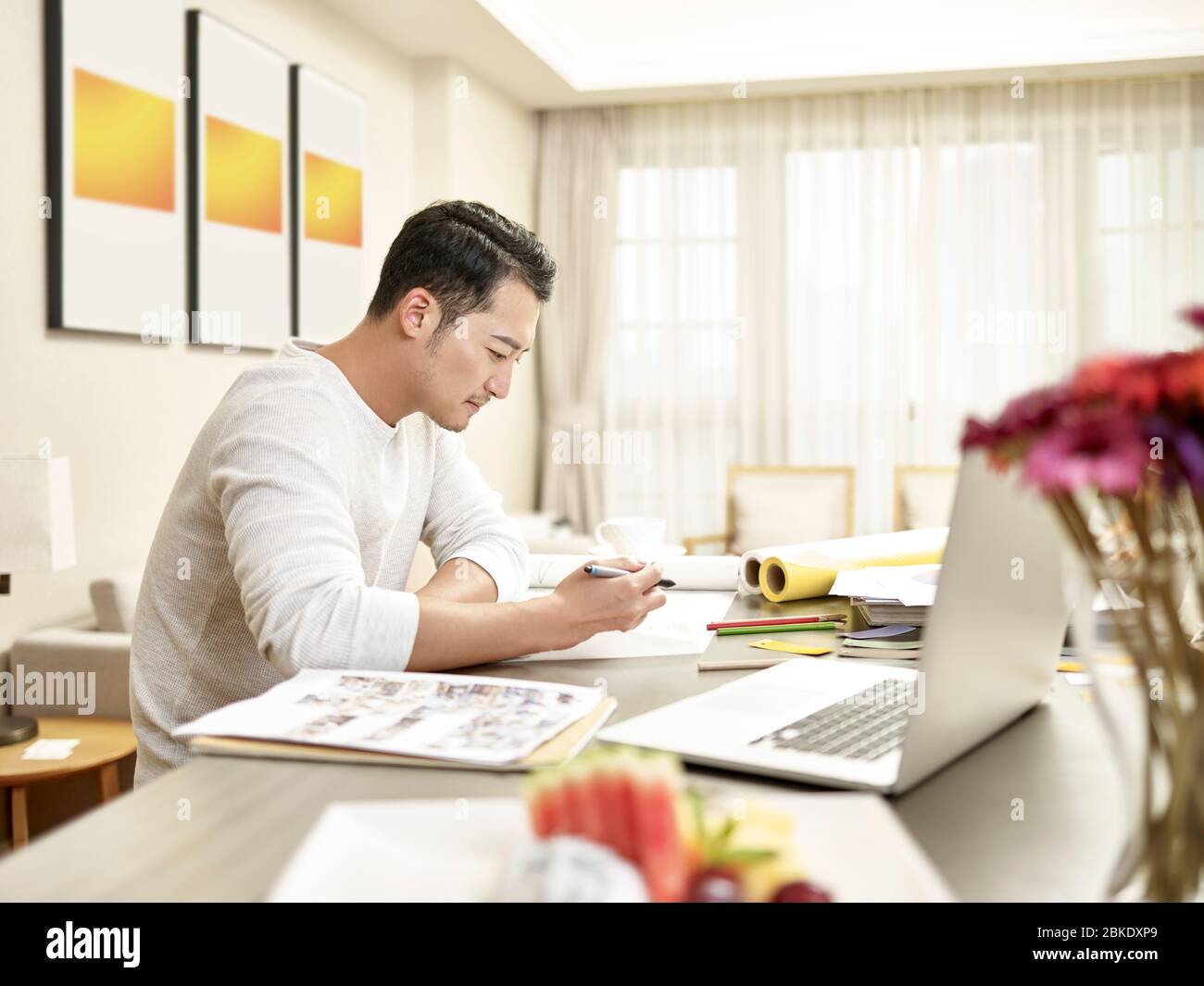 giovane asiatico uomo design professionale seduta al banco di lavoro da casa (grafica in background modificata digitalmente) Foto Stock