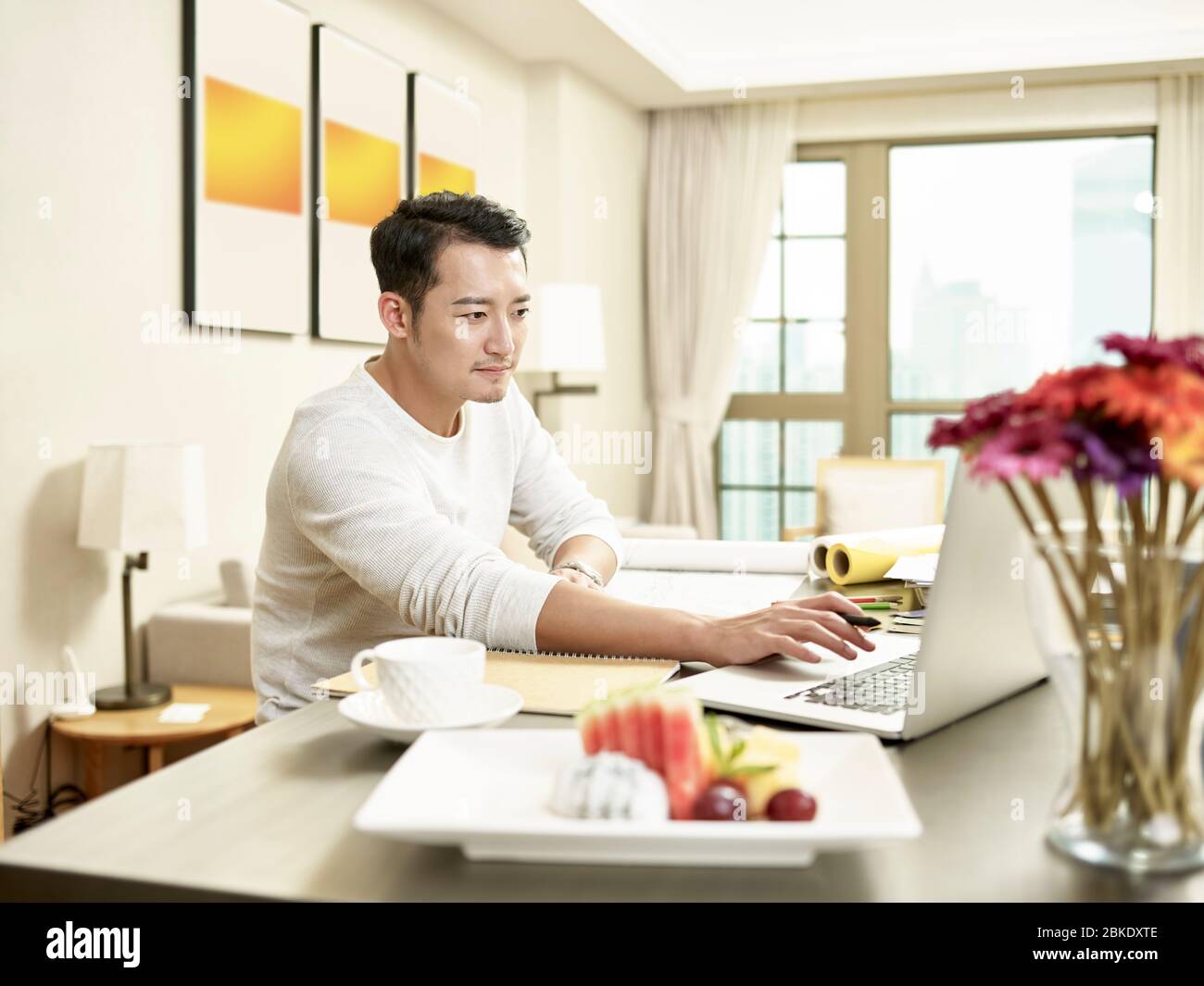 giovane uomo d'affari asiatico che lavora da casa seduto al bancone della cucina guardando il computer portatile (grafica in background modificata digitalmente) Foto Stock