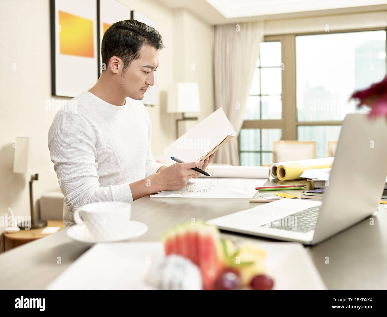 giovane asiatico uomo design professionale di lavoro da casa seduta in cucina guardando le note (grafica in background alterato digitalmente) Foto Stock