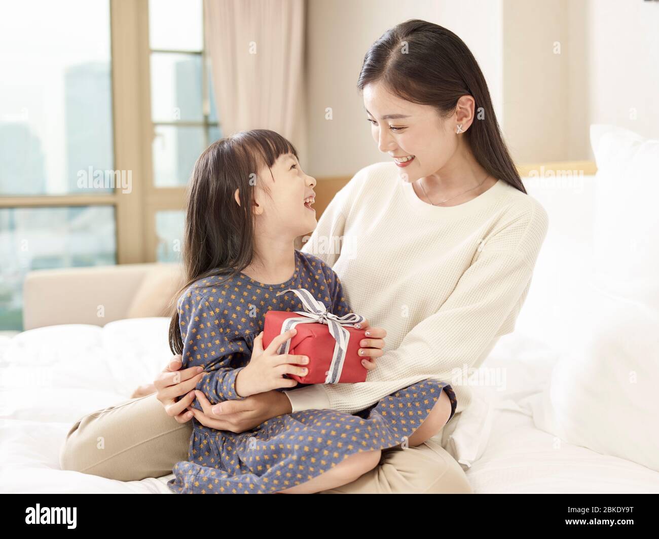 felice piccola ragazza asiatica seduta in braccia di giovane madre amorevole tenendo una scatola avvolta di regalo Foto Stock