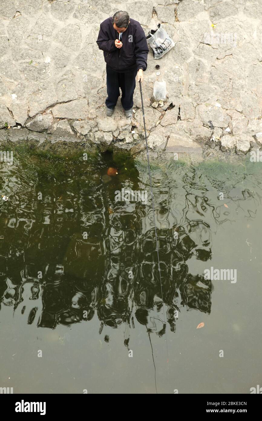 Vecchio che accende una sigaretta mentre tiene la canna da pesca mentre aspetta che il pesce sia agganciato. Pechino, Cina Foto Stock