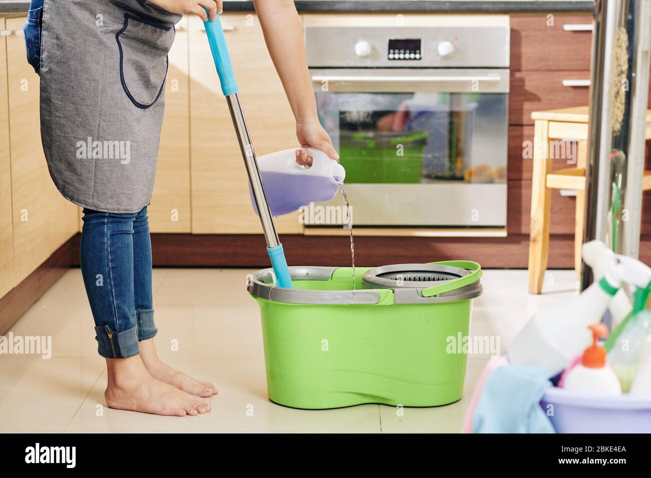 Casalinga in grembiule versando il liquido di pulizia del pavimento con profumo di lavanda in secchio con mop Foto Stock