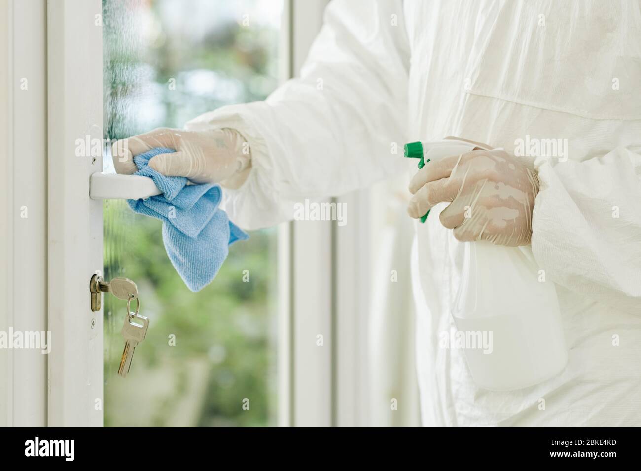 Immagine ravvicinata del tecnico in tuta protettiva e guanti che puliscono la maniglia della porta con spray chimici per uccidere i batteri e prevenire la diffusione delle malattie Foto Stock