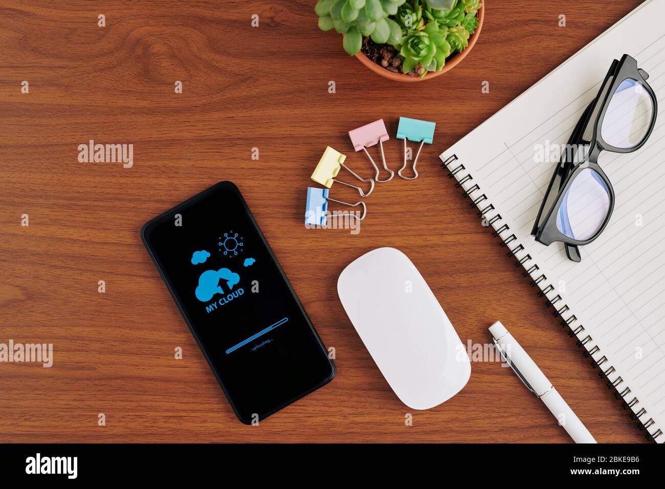 Smartphone con caricamento dei file sul servizio cloud tramite applicazione accanto a clip cartacee, libri di testo e mouse wireless sul tavolo dell'ufficio Foto Stock