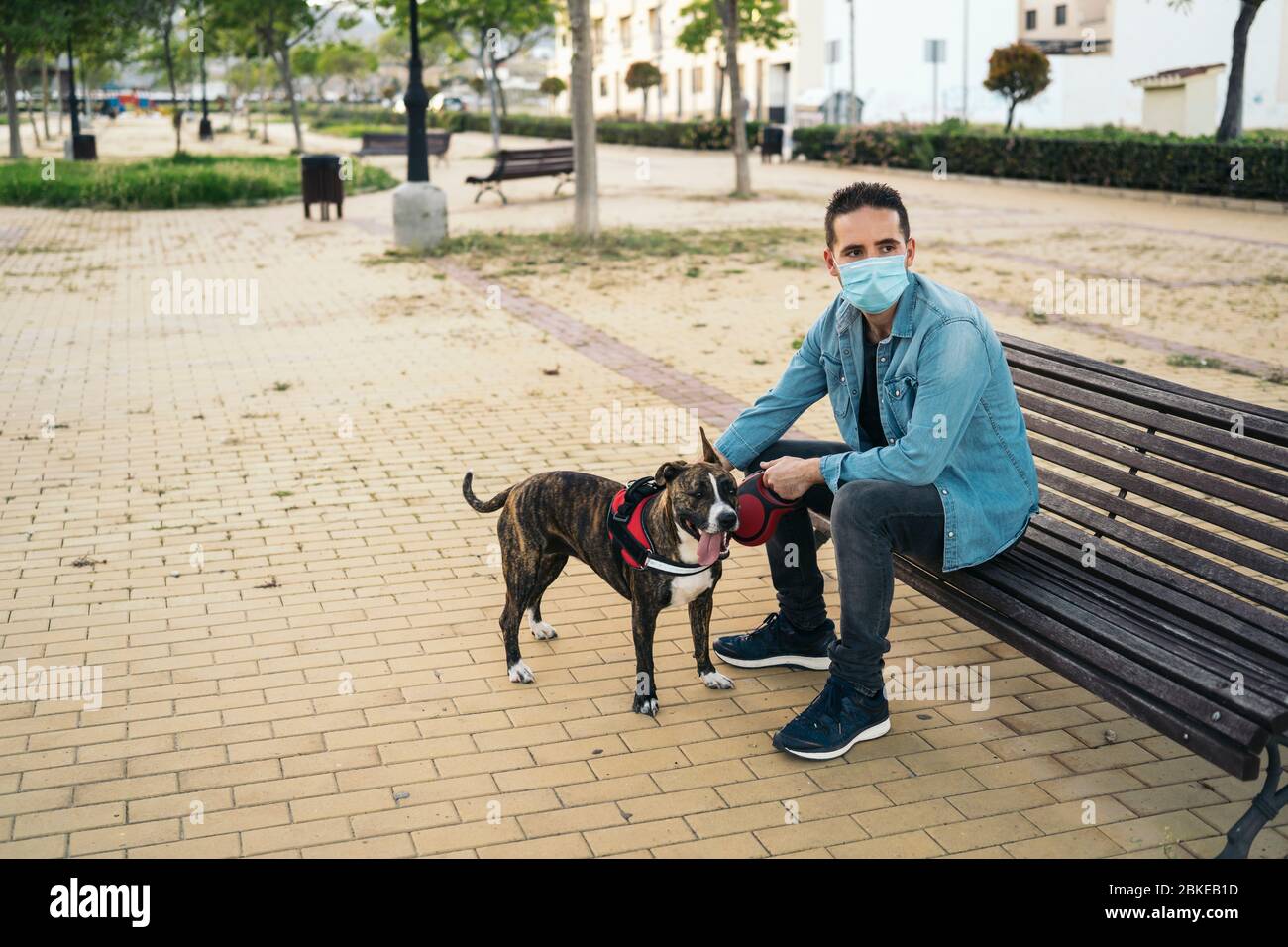 Giovane uomo in maschera medica seduto su una panchina nel parco con il suo cane. Covid-19 coronavirus pandemico. Vita attiva in quarantena nell'epidemia di Corona. Foto Stock