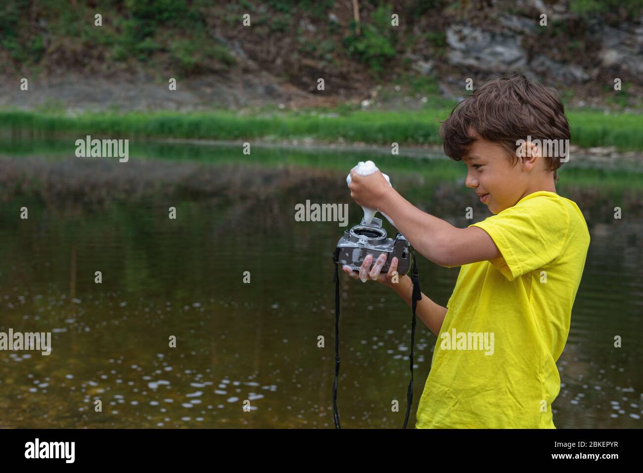 9 anni ragazzo in piedi in acqua, lava fotocamera digitale. Infanzia, prank infantile, macchina fotografica di pulizia sbagliata. Misure preventive per la diffusione dei virus. Foto Stock