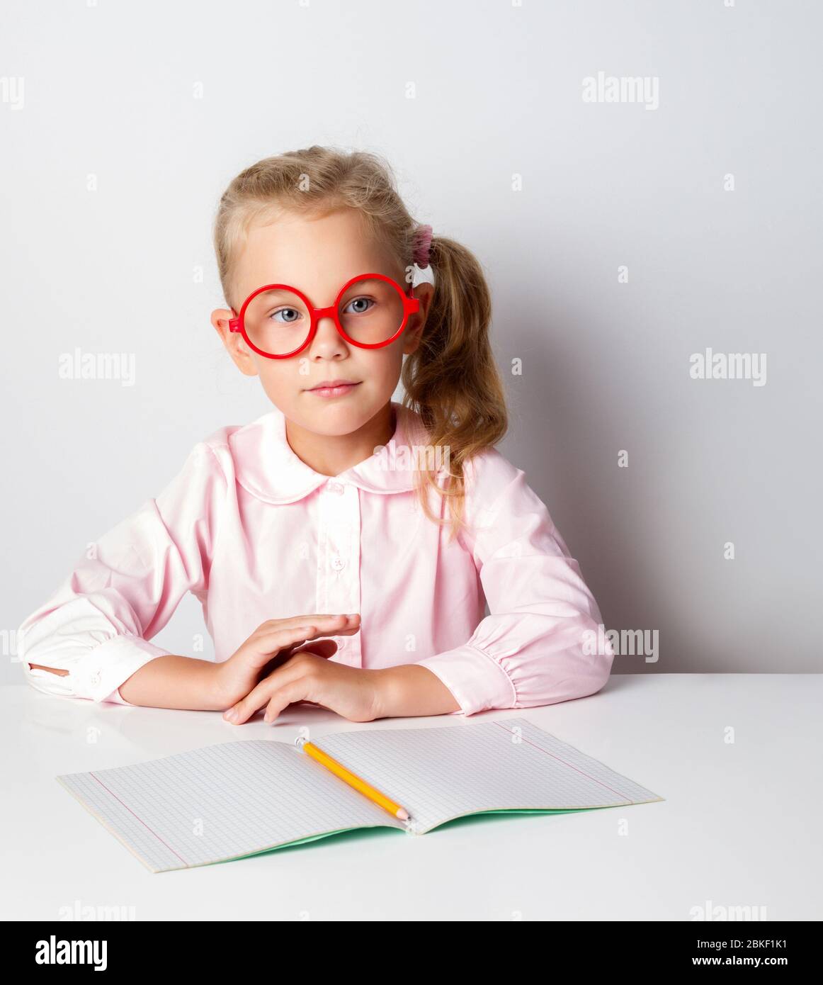 Piccola bionda in occhiali e blusa rosa. Seduta a tavola con copybook e matita su di esso. Posa isolata su bianco Foto Stock