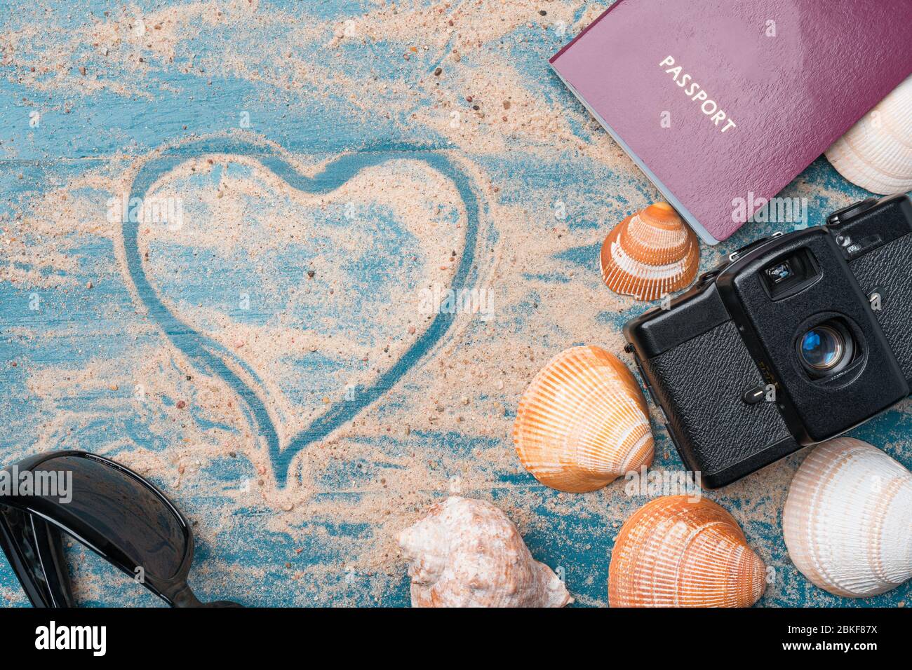 Passaporto, macchina fotografica vintage, occhiali da sole e conchiglie su uno sfondo di legno blu con sabbia dorata e forma di cuore. Viaggio estivo amore abstrac Foto Stock