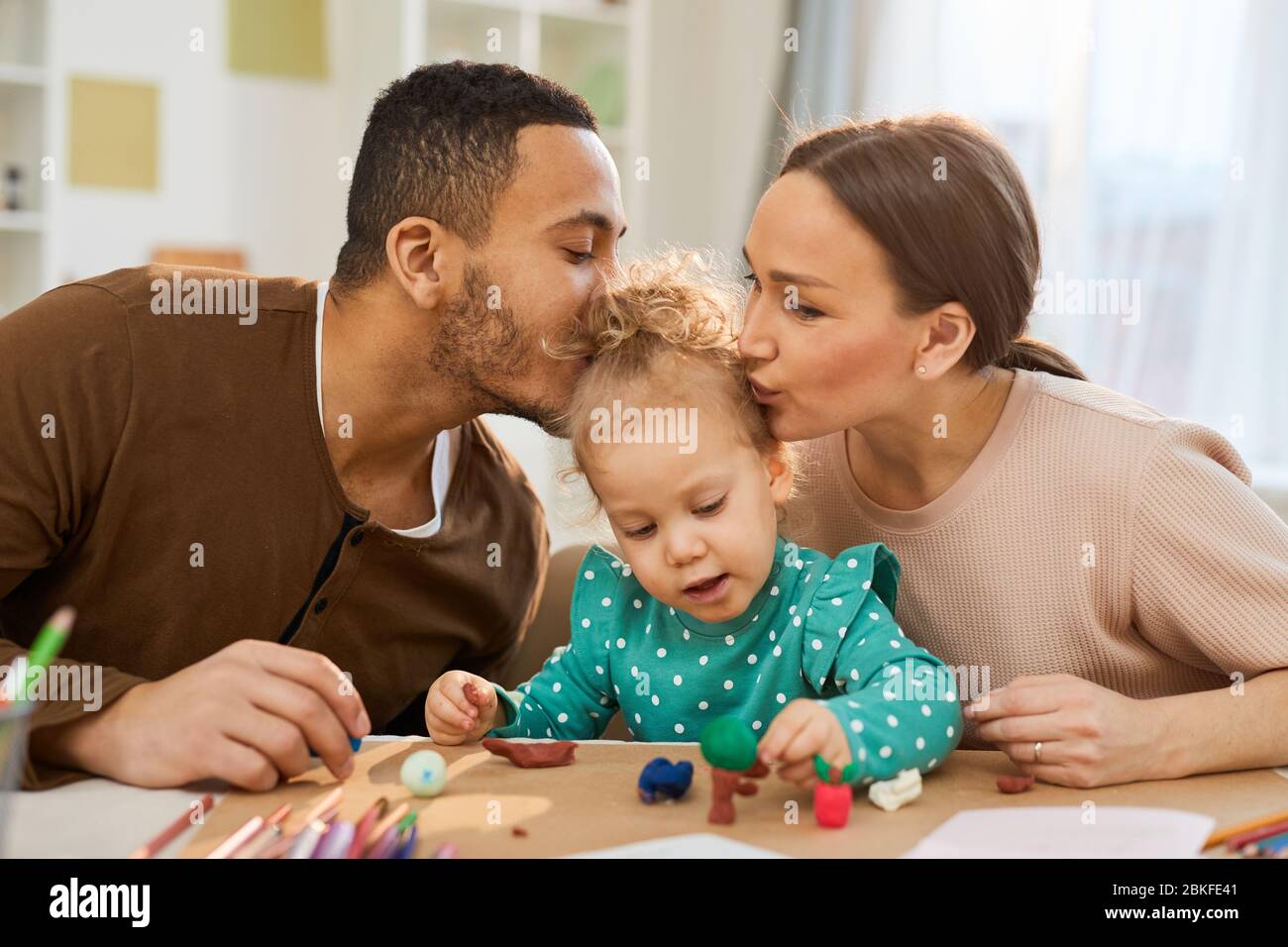 Padre e madre amorosi baciare la loro bambina cute mentre sta facendo i modelli colorati di pasta gioco, colpo orizzontale Foto Stock