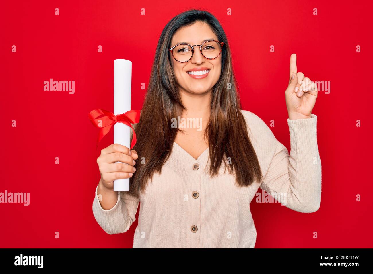 Giovane donna intelligente ispanica che indossa occhiali che tengono il grado universitario su sfondo rosso sorpreso con un'idea o domanda che punta il dito con felice Foto Stock