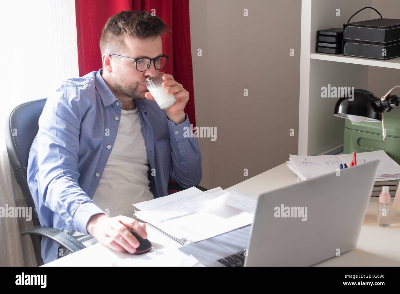 Uomo caucasico che lavora a casa sul computer portatile leggendo documenti e bevendo latte. Foto Stock