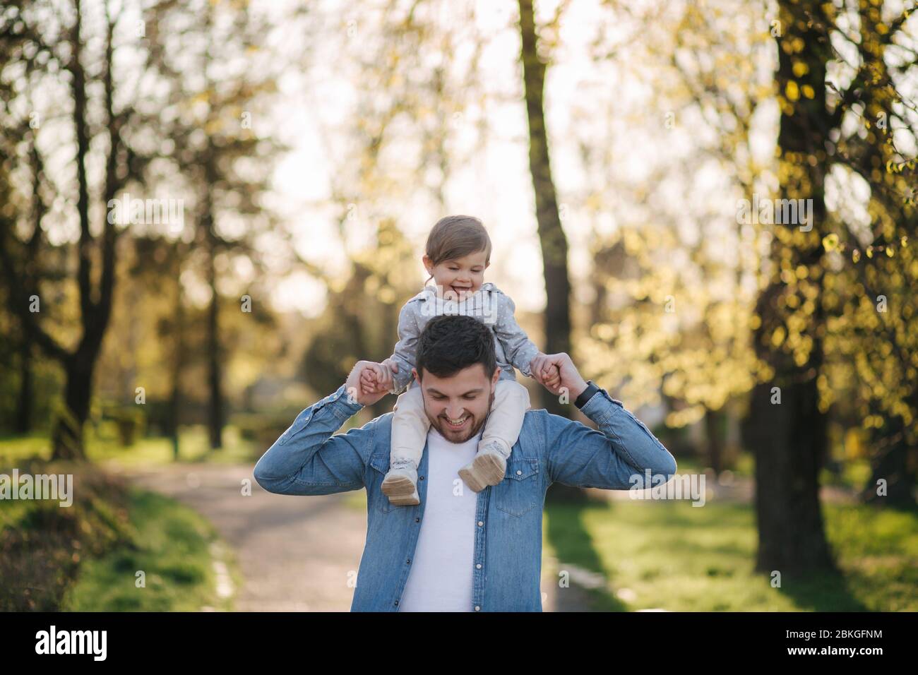 Adorabile figlia seduta sul collo del papà e ridendo. Giovane padre cammina con la sua bambina carina nel parco Foto Stock