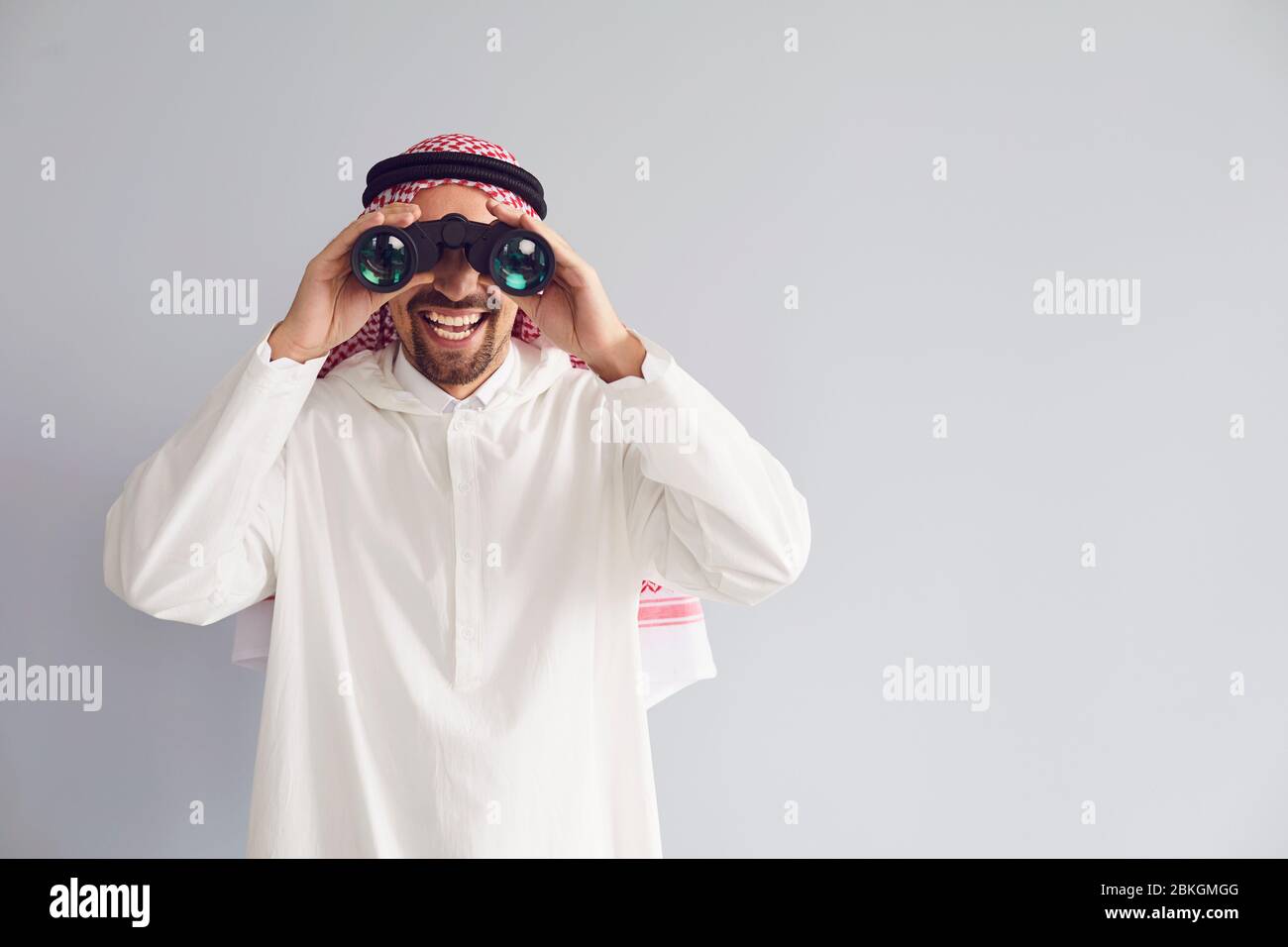 Uomo arabo che guarda attraverso binocoli sorridendo su uno sfondo grigio. Foto Stock