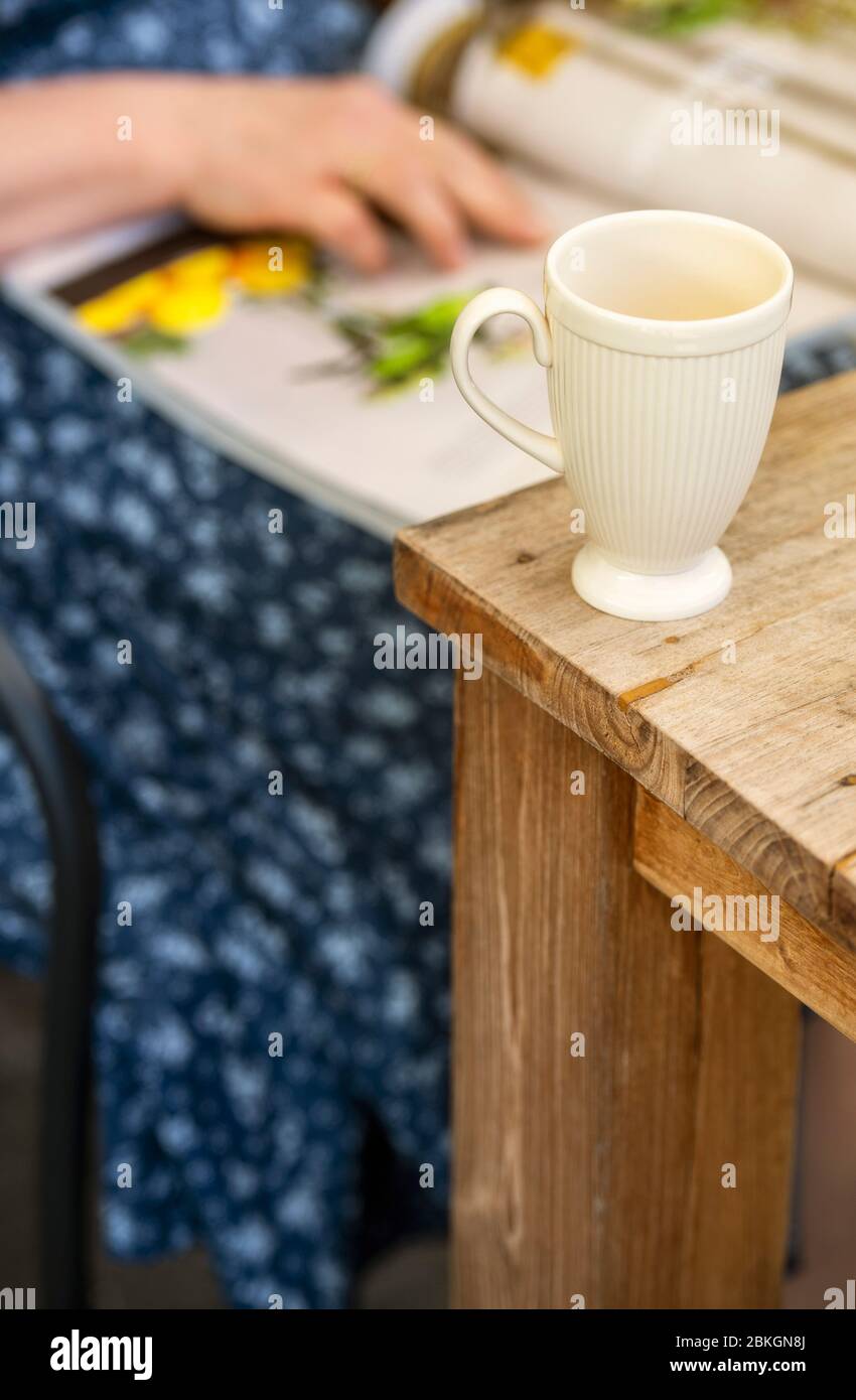 Donna che legge una rivista all'aperto con una tazza vuota vicino su un tavolo da giardino Foto Stock