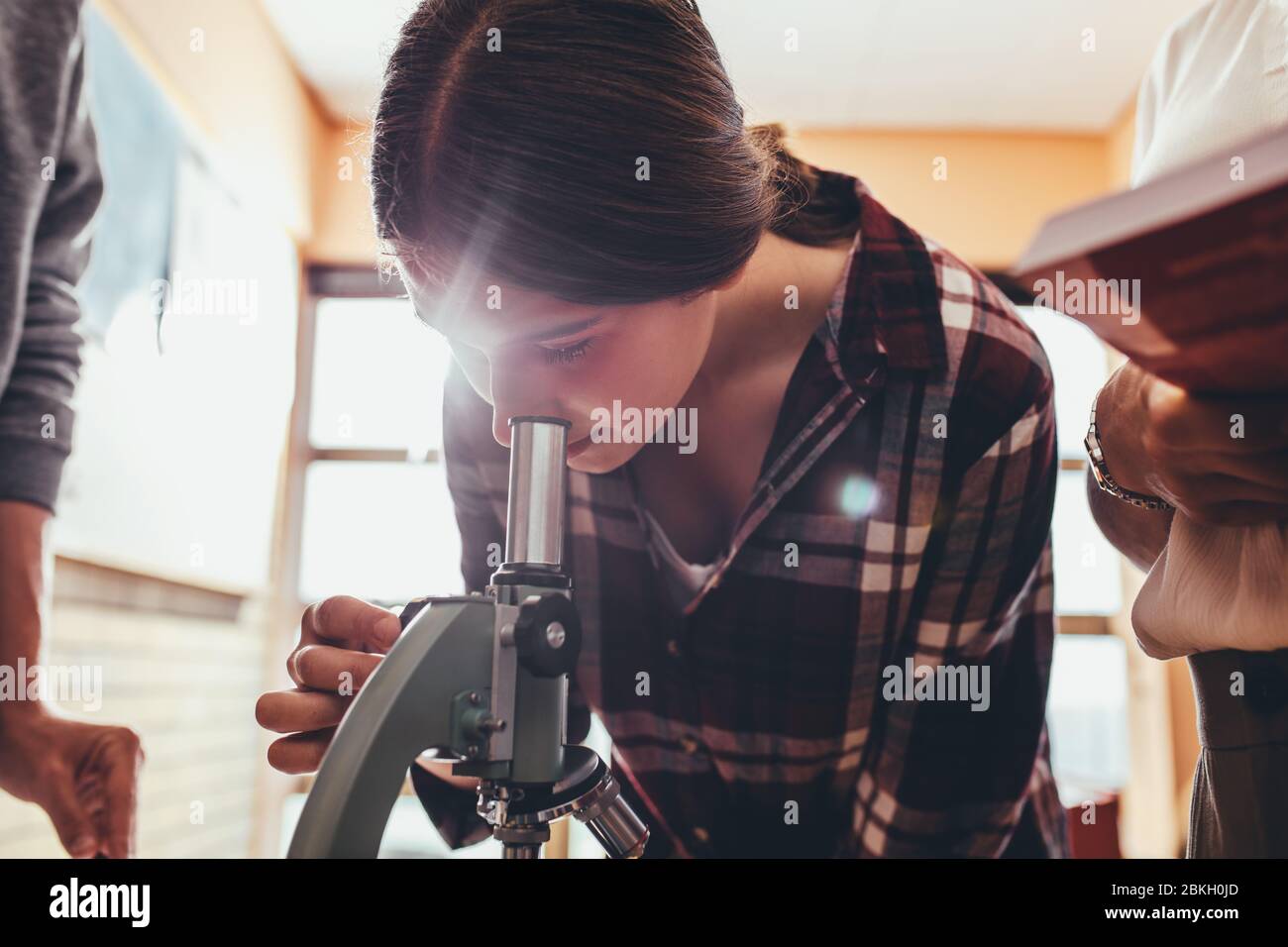 Scuola ragazza in classe di scienza utilizzando un microscopio con insegnante e compagno di classe in piedi. Studente che guarda attraverso il microscopio in classe di biologia. Foto Stock