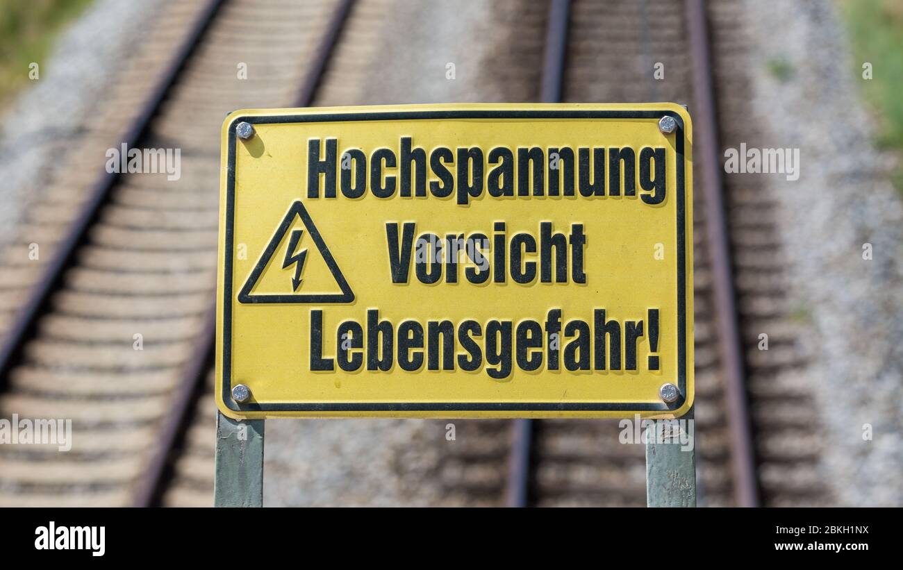 Segno giallo con la scritta "Hochspannung, Vorsicht, Lebensgefahr" (alta tensione, attenzione, pericolo per la vita). Formato quadrato. Sullo sfondo binari ferroviari Foto Stock