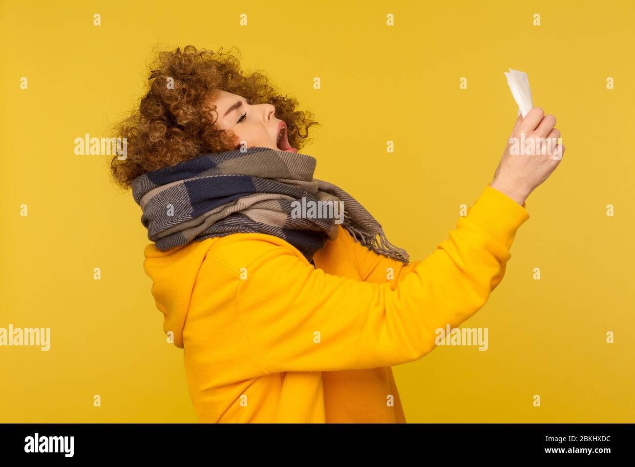 Influenza stagionale. Vista laterale della donna malata di influenza con capelli ricci morbidi, avvolti in sciarpa calda, starnuti in tovagliolo, sensazione di malessere, tosse e blowin Foto Stock