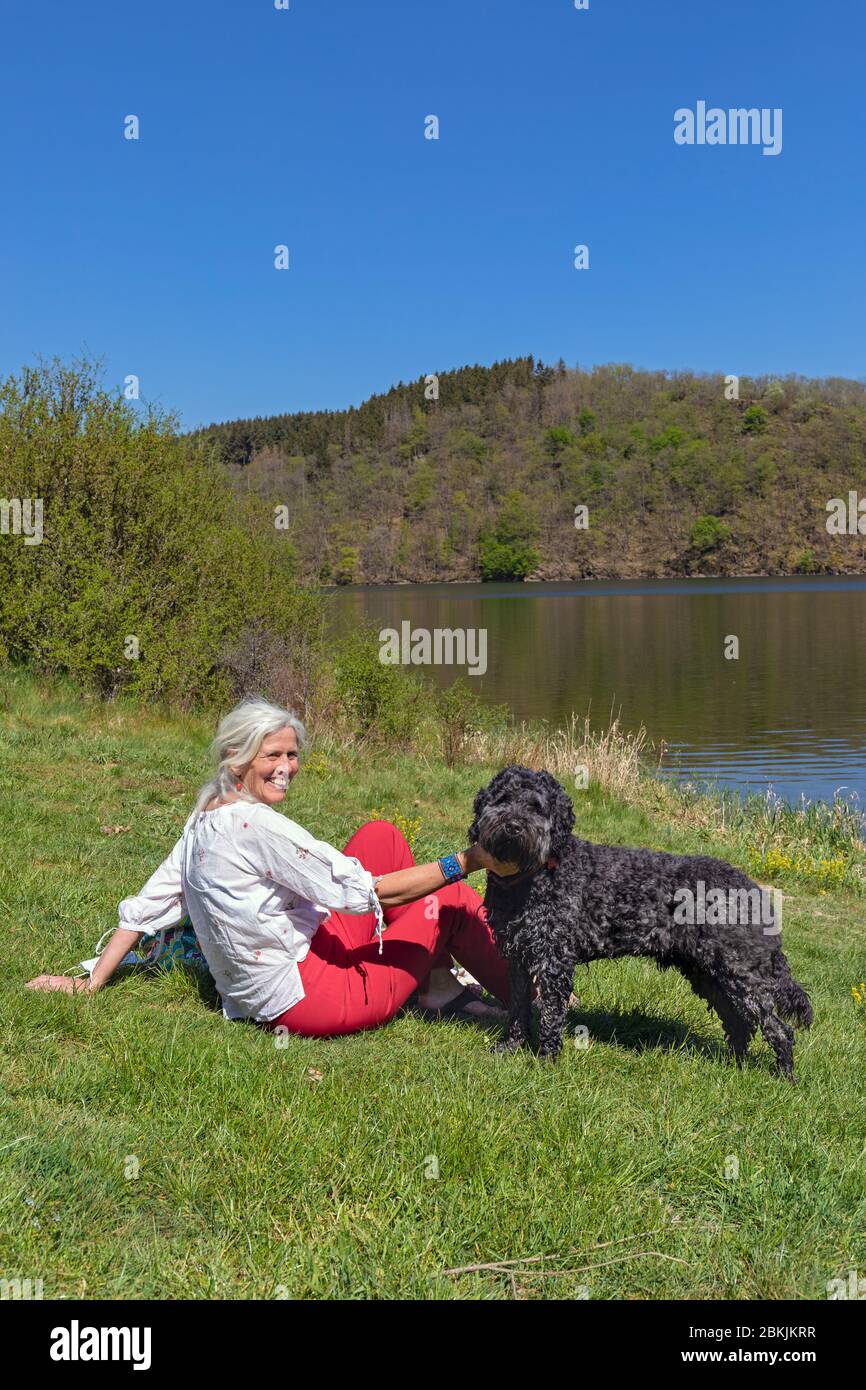 Europa, Lussemburgo, Insenborn, attraente donna anziana seduta sulla riva del Lac Sûre con il suo cane d'acqua portoghese Foto Stock