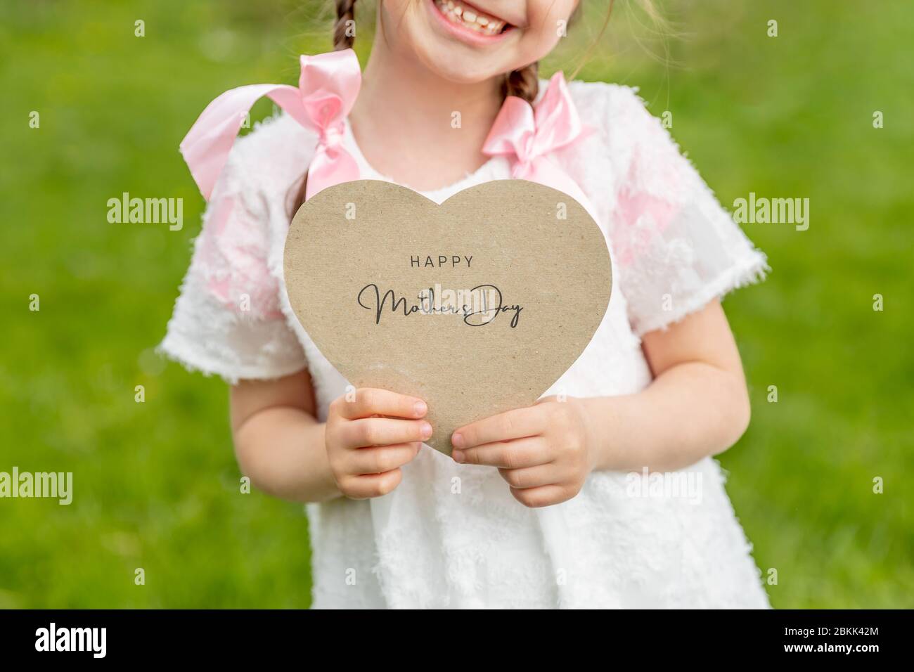 Felice Festa della mamma scritta su una carta a forma di cuore tenuta da una ragazza su sfondo d'erba Foto Stock
