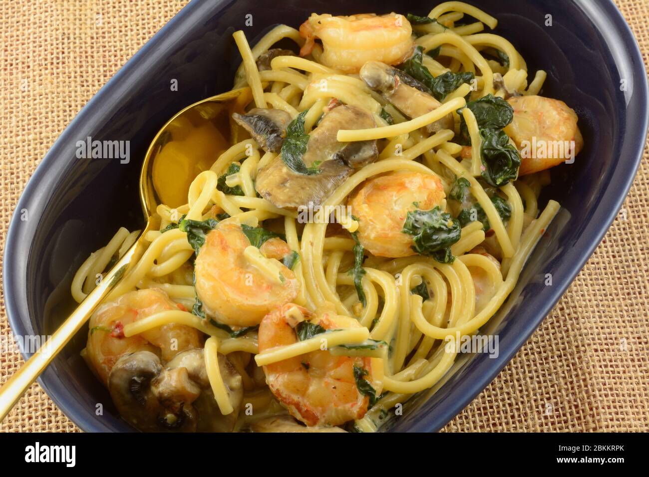 Primo piano di gamberi e pasta cena con spinaci e funghi in salsa crema leggera in ciotola blu con cucchiaio dorato Foto Stock