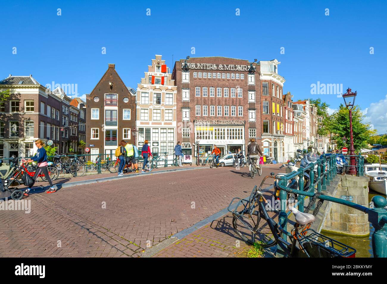 Biciclette parcheggiate lungo un canale vicino alla Chiesa Westerkerk, mentre i turisti e la gente del posto si godono i negozi e i ponti nel centro storico di Amsterdam Nether Foto Stock