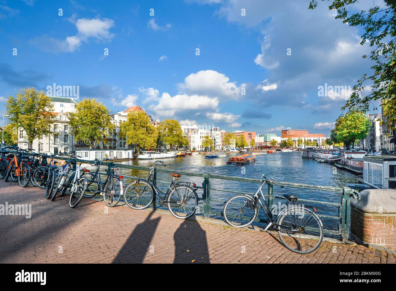 Le biciclette si parcheggiano lungo un ponte su uno dei canali più grandi del centro storico di Amsterdam mentre una barca turistica viaggia in lontananza Foto Stock