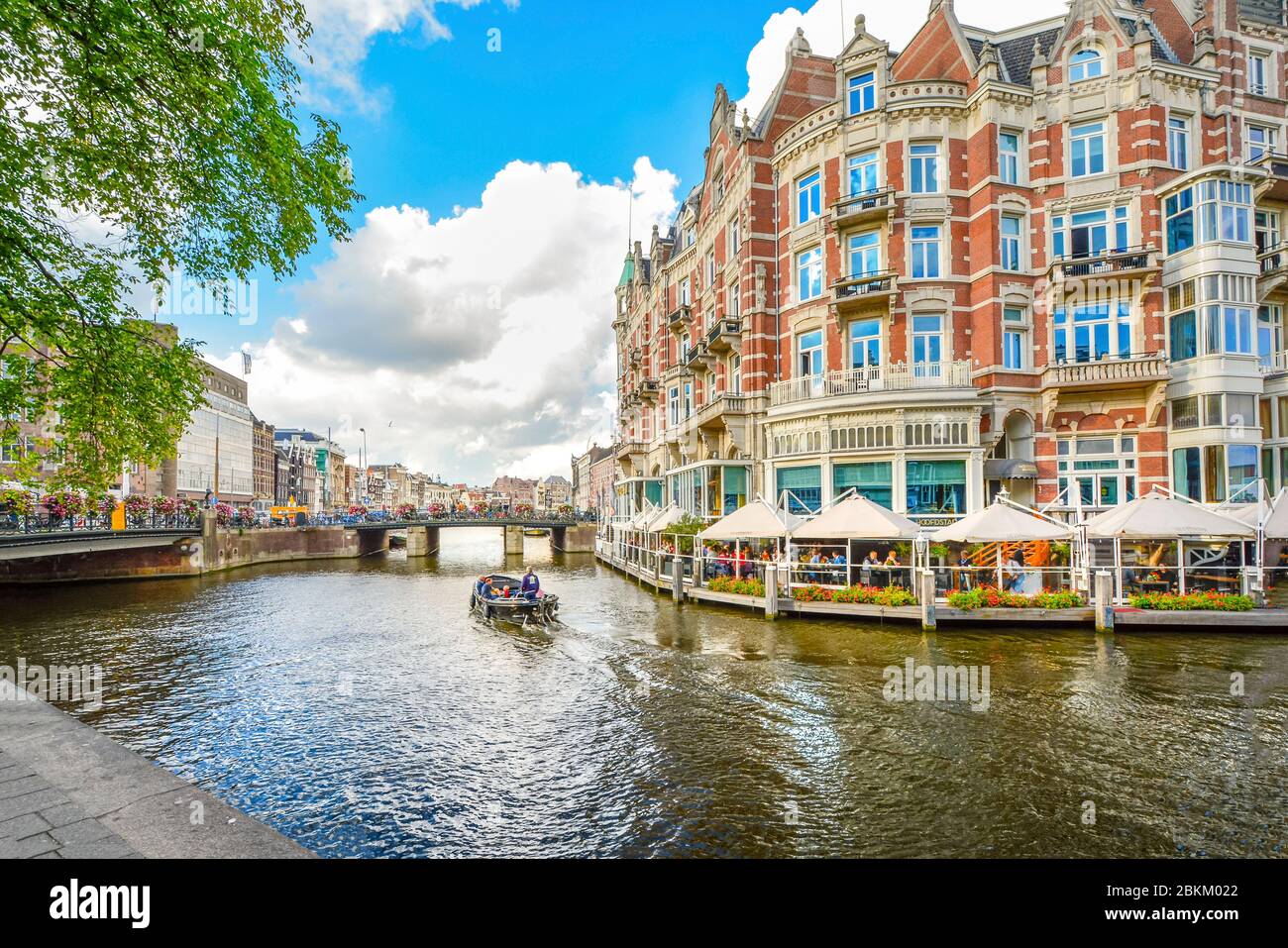 Una barca scivola passato a waterfront hotel e il cafe verso un ponte su uno dei canali principali della cittã , vicino al quartiere dei musei di Amsterdam, Paesi Bassi. Foto Stock