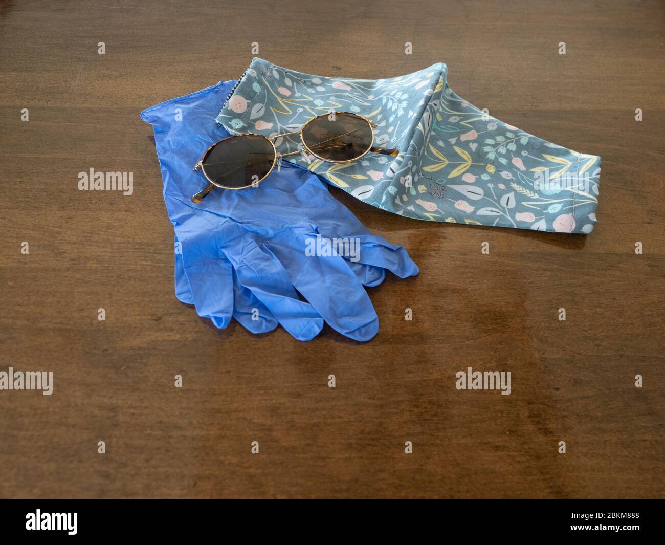 Un paio di guanti monouso blu, occhiali da sole e una maschera in tessuto floreale grigio artigianale su un tavolo di legno. Fotografato con messa a fuoco selettiva. Imag Foto Stock