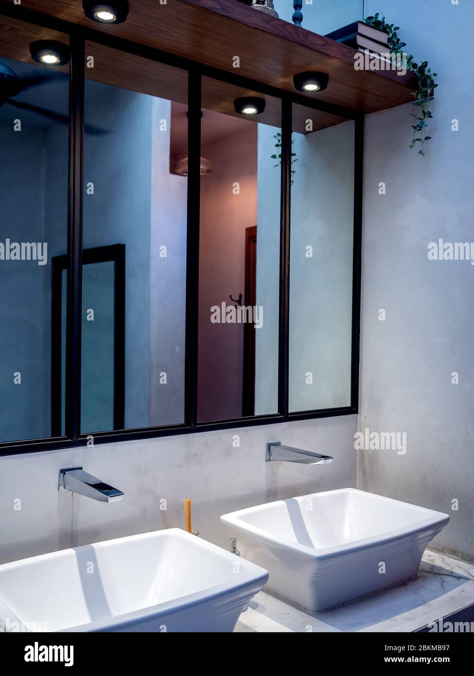 Lavabo e rubinetto bianchi moderni con specchio alto, downlight e decorazione di scaffali in legno su pareti in cemento in bagno, stile verticale. Foto Stock