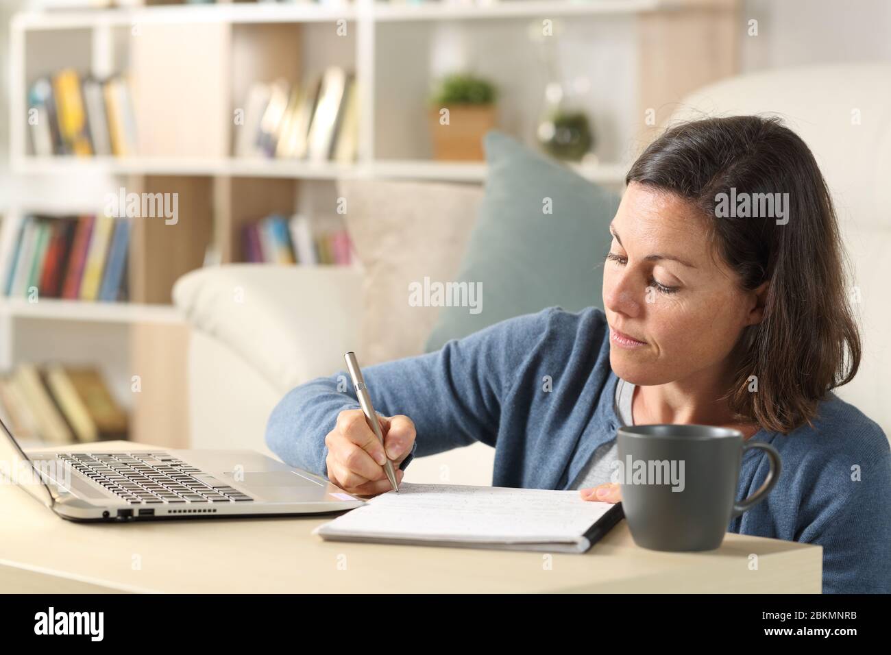 Donna adulta con notebook che scrive appunti sul notebook seduto sul pavimento su un tavolino a casa Foto Stock