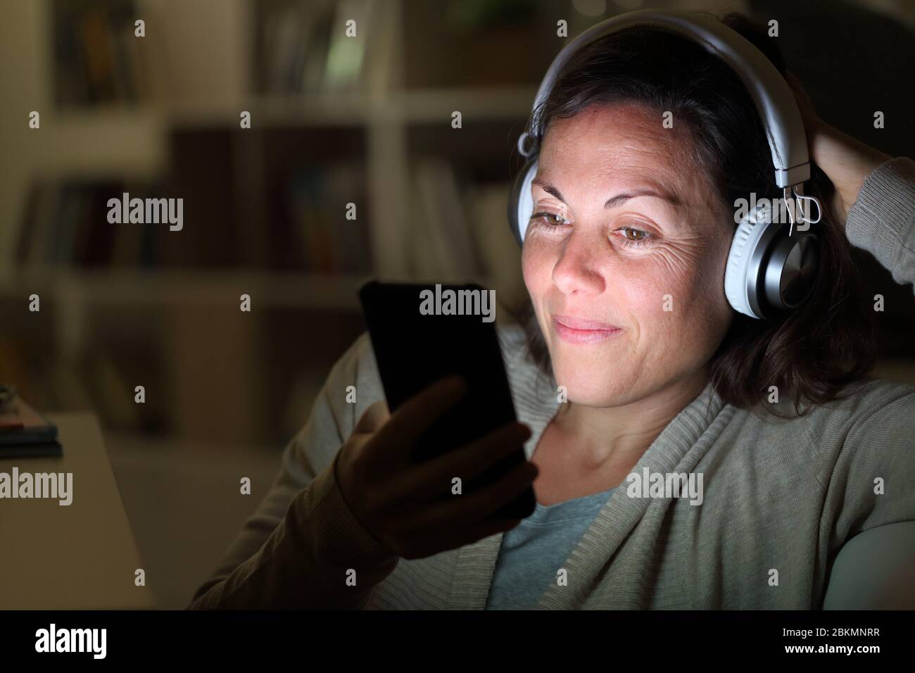 Una donna adulta felice con cuffie ascolta la musica sullo smartphone con schermo illuminato seduto a casa di notte Foto Stock