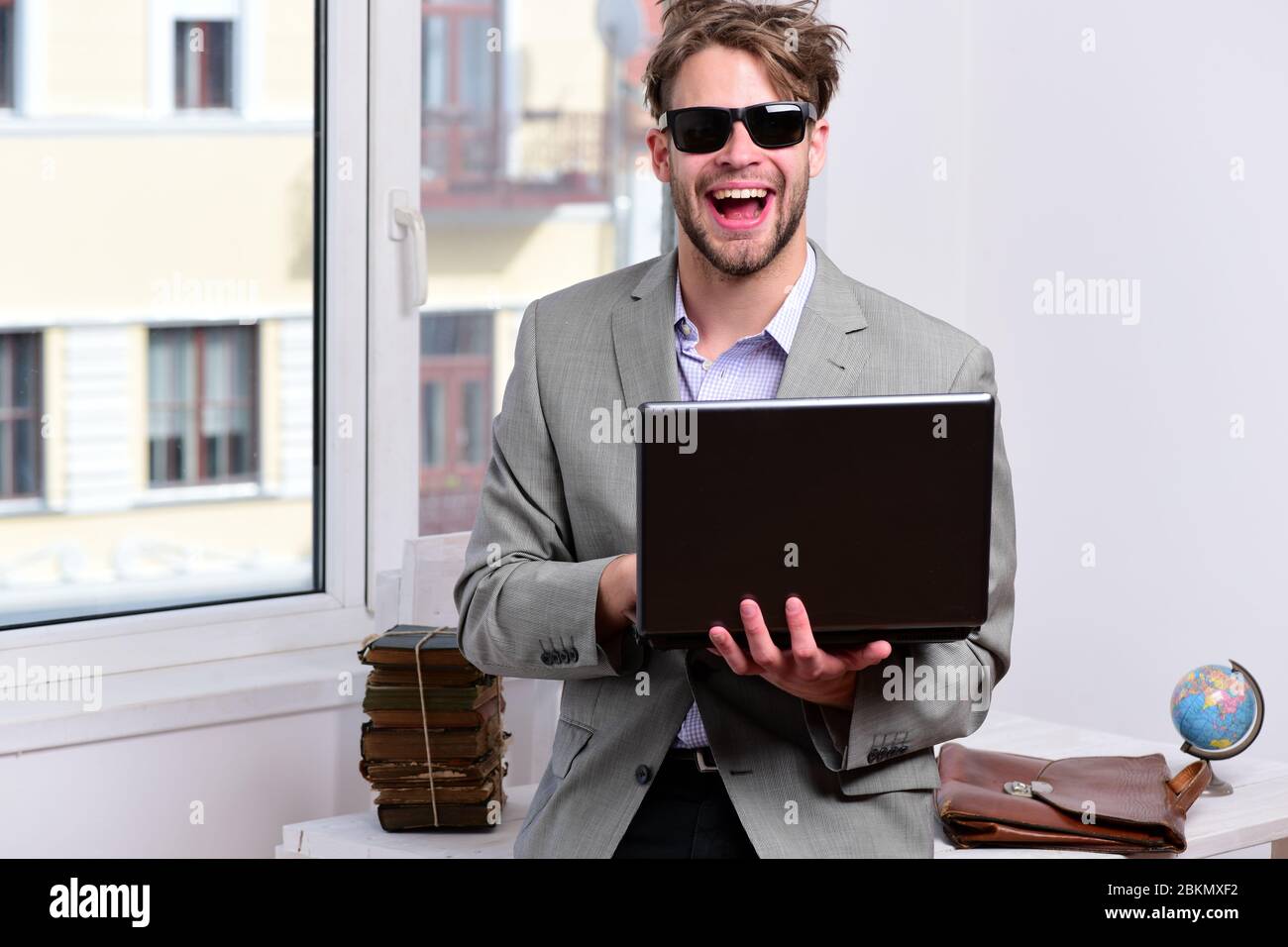 Uomo o manager con setole e viso allegro in occhiali da sole. Uomo d'affari  con laptop e mucchio di libri sullo sfondo della finestra dell'ufficio. Il  ragazzo fresco si siede sul tavolo
