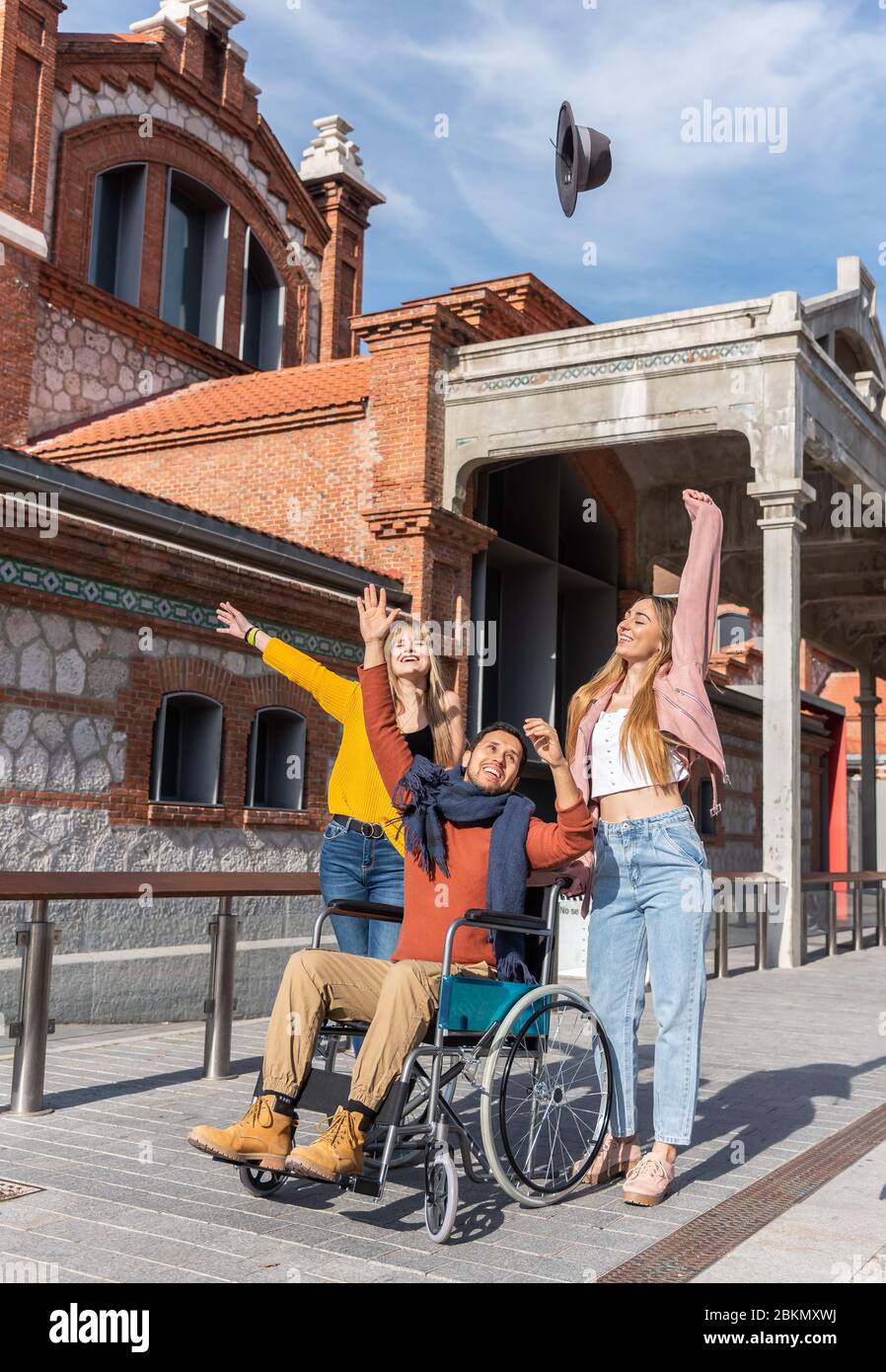 Paraplegico giovane latino-americano uomo in una sedia a rotelle accanto a due giovani ragazze caucasiche felice buttare un cappello in aria mentre si cammina per la strada Foto Stock