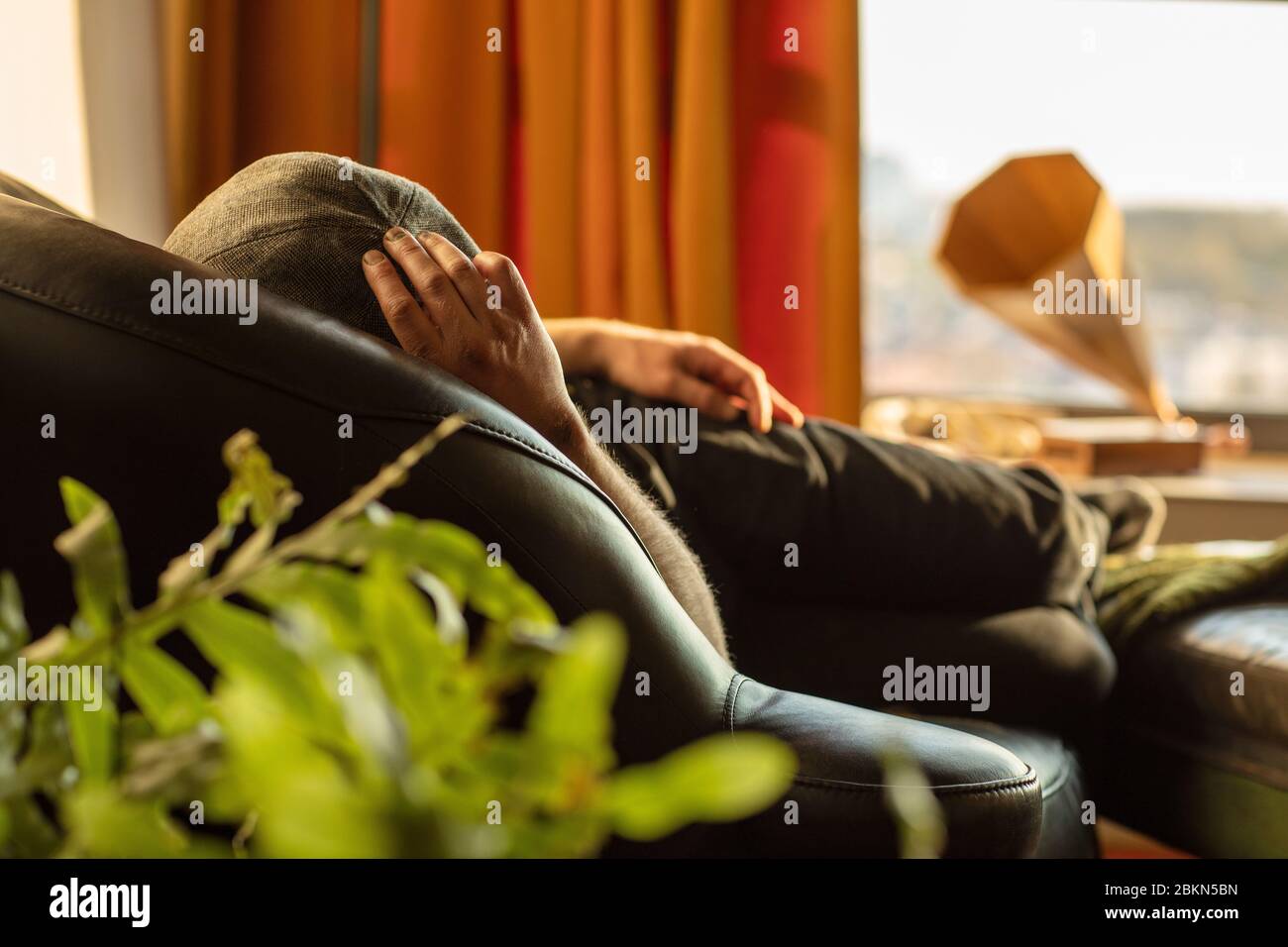 Un uomo che indossa una cuffia piatta rilassarsi e prendere un pisolino mentre si rimane all'interno sul divano una giornata di sole in un interno retrò, vintage, eclettico stile con o. Foto Stock