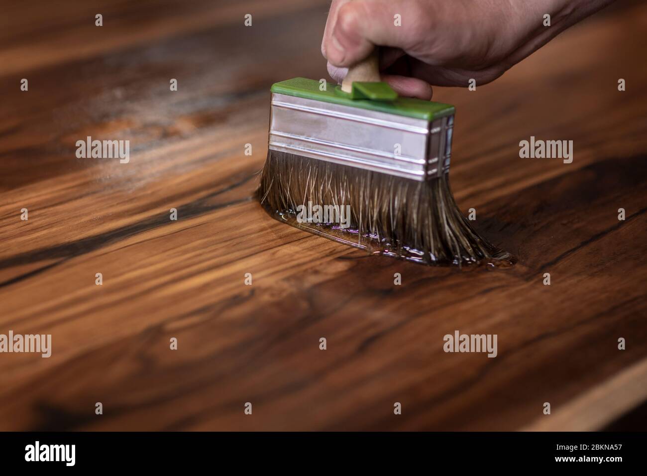 Mano di un carpentiere designer che oliando un autentico tavolo fatto da sé con una grande spazzola in un ambiente di officina. Artigiano rifinendo le lastre di legno di teak con Foto Stock