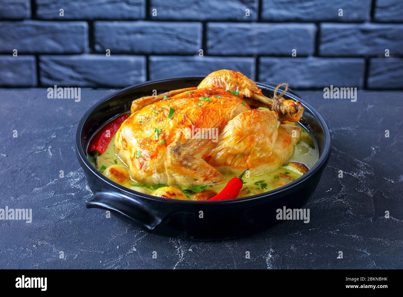 primo piano di pollo arrosto al cocco con patata in una brasiera in ceramica nera su un tavolo di cemento con un muro di mattoni sullo sfondo, horizta Foto Stock