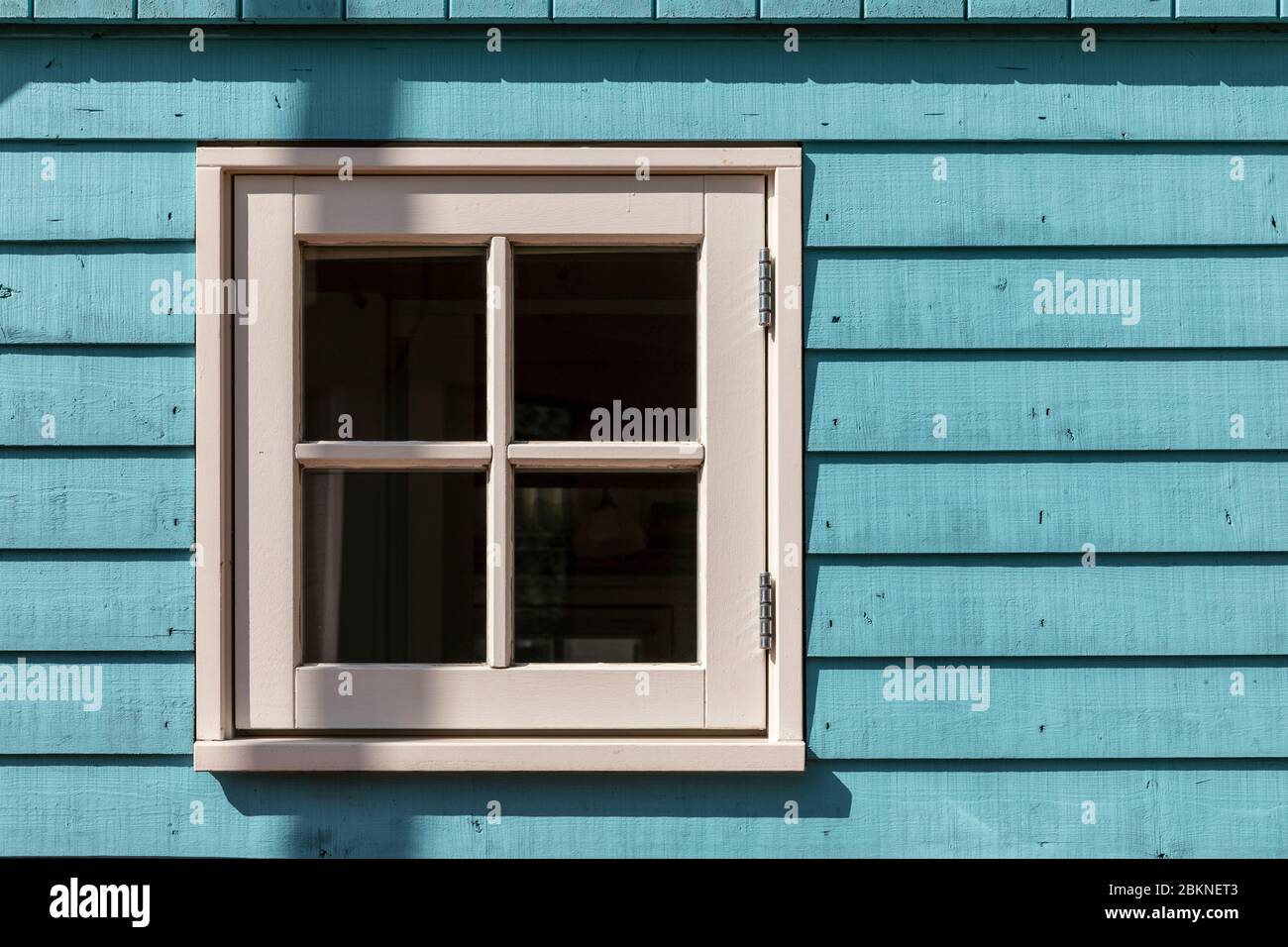 Una piccola finestra di una piccola casa con una cornice bianca e una facciata di legno duro di colore blu chiaro composta da pannelli in una giornata di sole. Scatto orizzontale Foto Stock