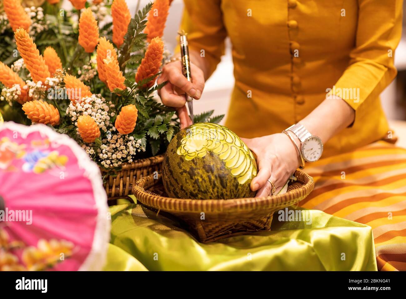 Una donna che intagliano un anguria circondata da verde e fiori d'arancio. Cibo decorato, a forma di disegno con molti dettagli. Tradizione tailandese Foto Stock