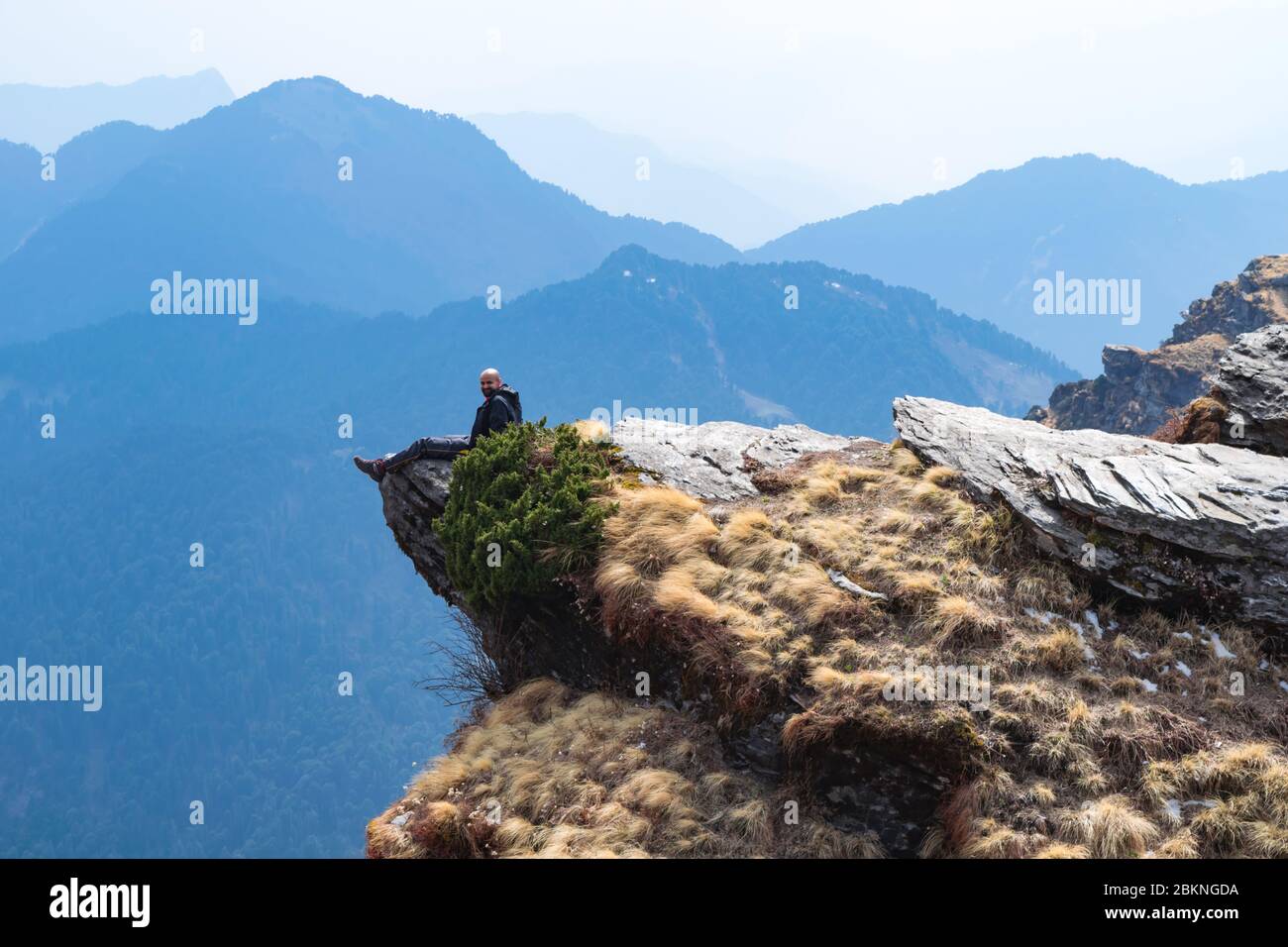 Giovane escursionista indiano calvo seduto al bordo di una scogliera di montagna con le gambe giù per la collina e godere della vista mozzafiato Foto Stock