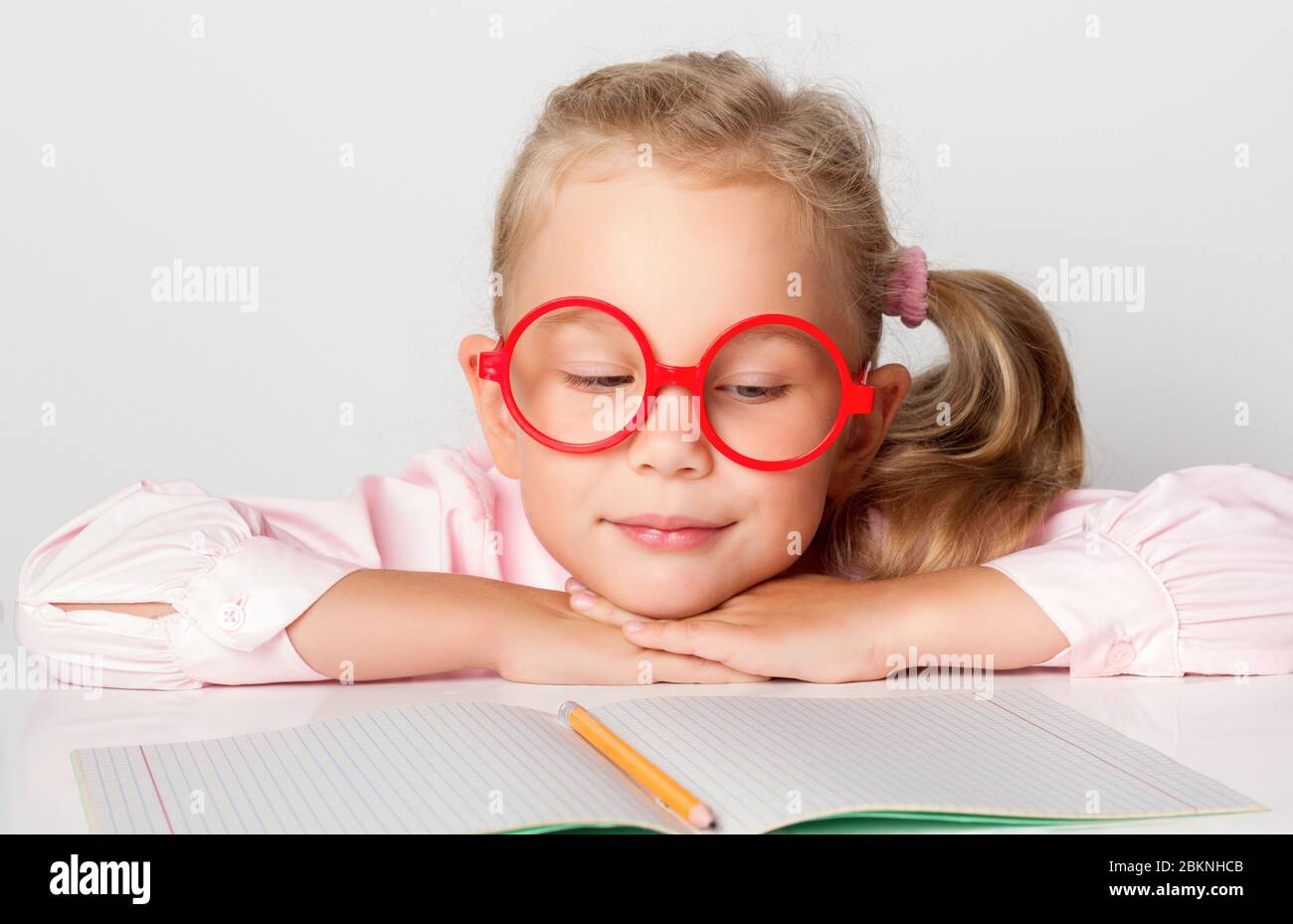 Bionda in occhiali rossi incorniciati, blusa rosa. Sorridente, appoggiato su una tavola con copybook e matita su di esso. Posa isolata su bianco Foto Stock