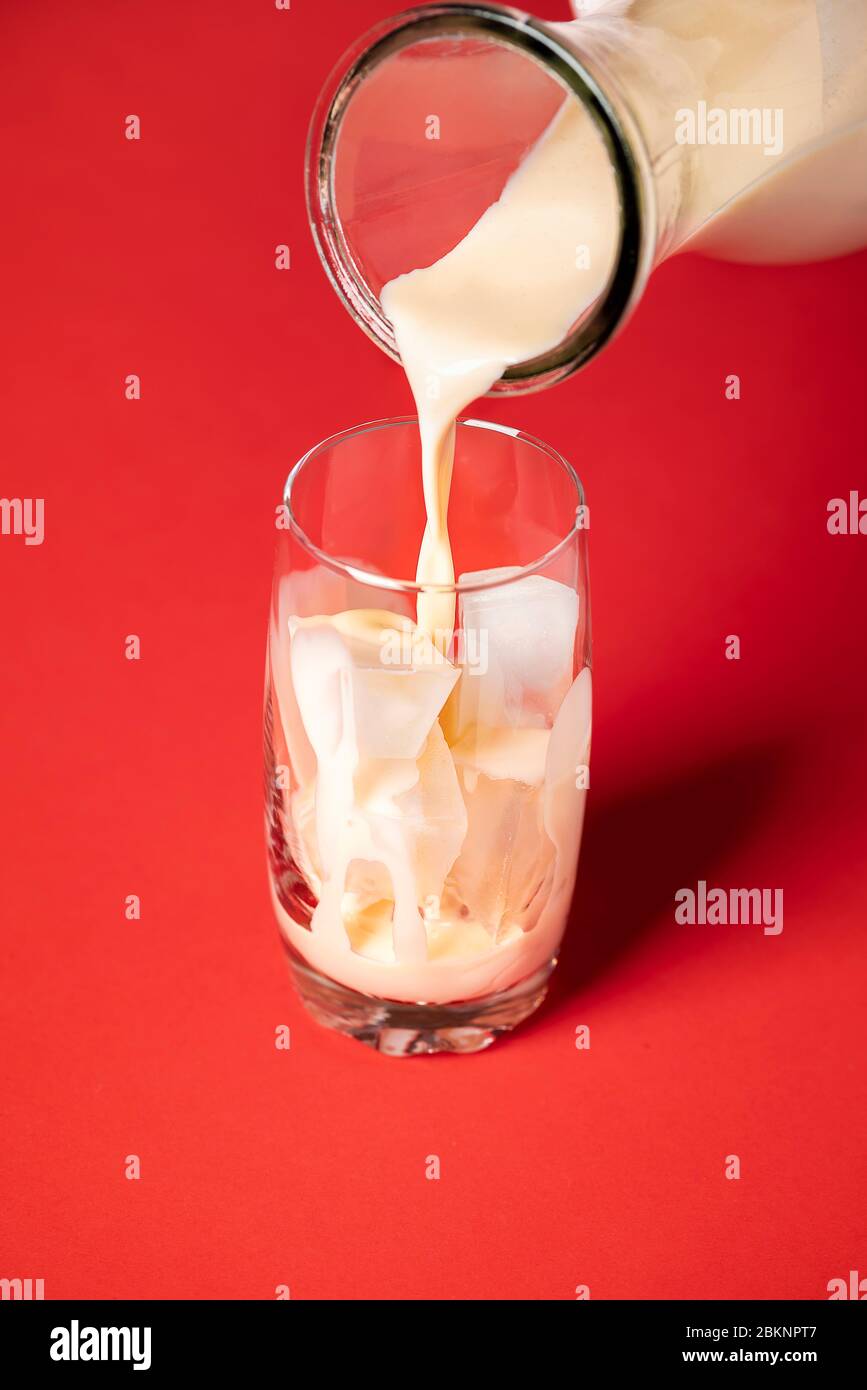 Fare horchata mocktail contesto. Versare il latte di horchata in un bicchiere con cubetti di ghiaccio su fondo rosso. Serve drink estivi messicani. Bevanda fatta in casa Foto Stock