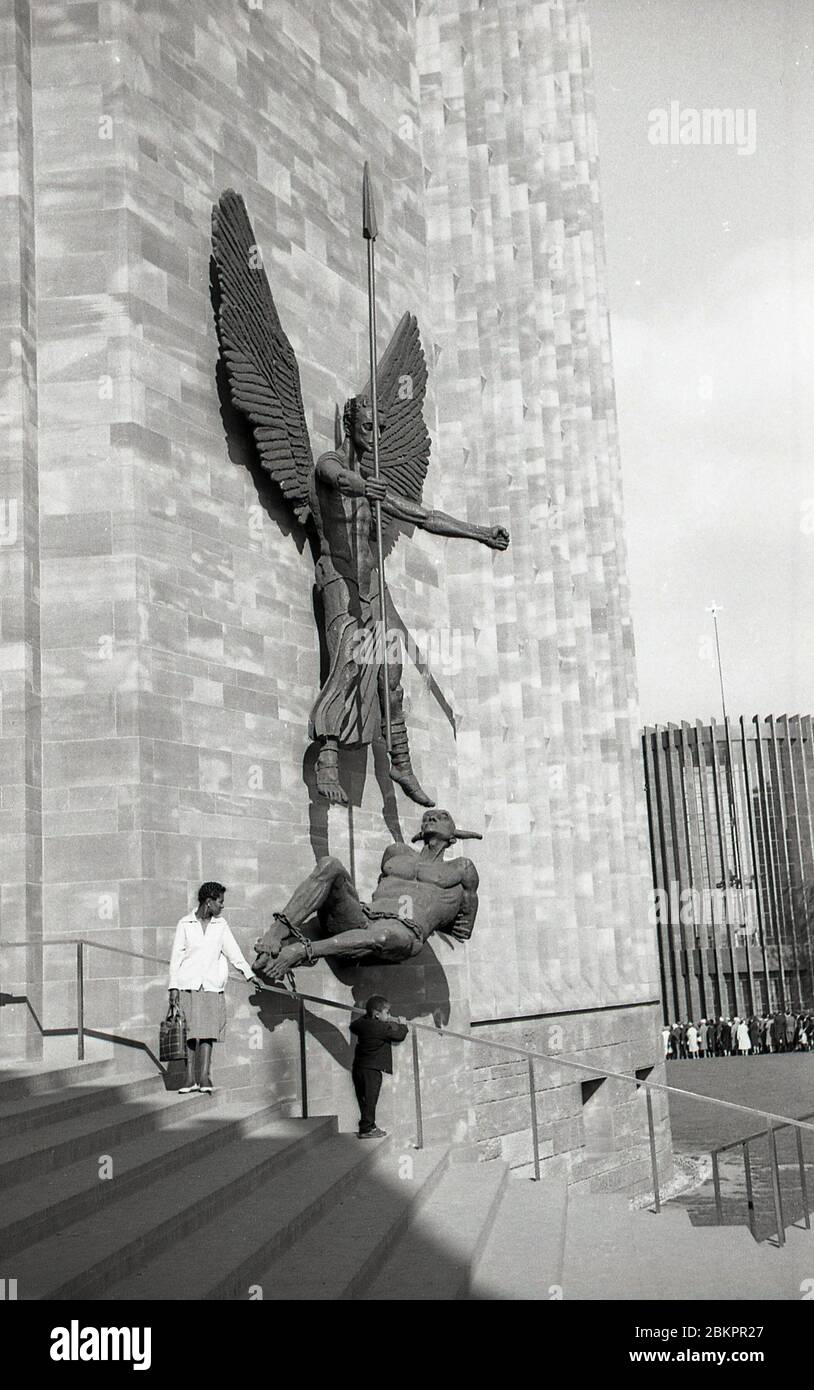 1962, quadro storico che mostra una signora e suo figlio che si trovano fuori dalla cattedrale di Coventry, Inghilterra. La scultura sul muro, di Jacob Epstein, denota la vittoria di San Michele sul Diavolo. Pesantemente bombardato durante la seconda guerra mondiale, nel maggio 1962 fu aperto un nuovo edificio 'modernista' in città, sul sito delle rovine della vecchia cattedrale. Foto Stock