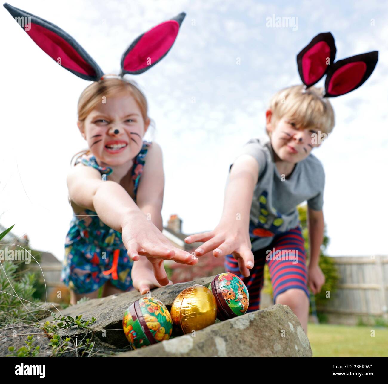 Un fratello e una sorella, indossando la pittura facciale e le orecchie conigliate fatte in casa, prendere parte a una caccia all'uovo di Pasqua la Domenica di Pasqua nel loro giardino. Foto Stock