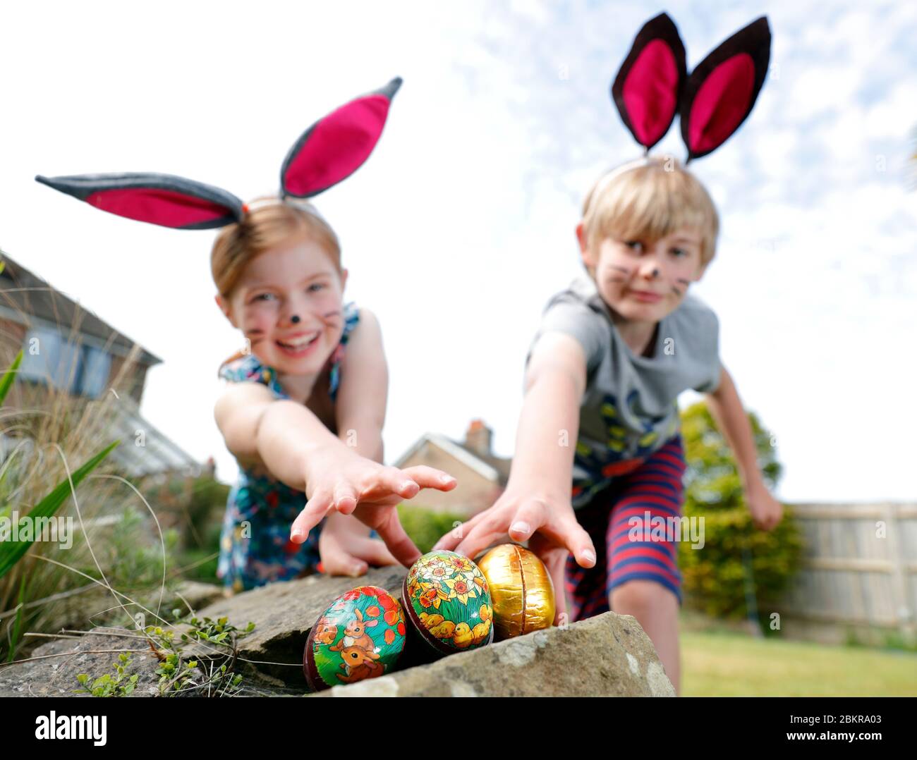 Un fratello e una sorella, indossando la pittura facciale e le orecchie conigliate fatte in casa, prendere parte a una caccia all'uovo di Pasqua la Domenica di Pasqua nel loro giardino. Foto Stock