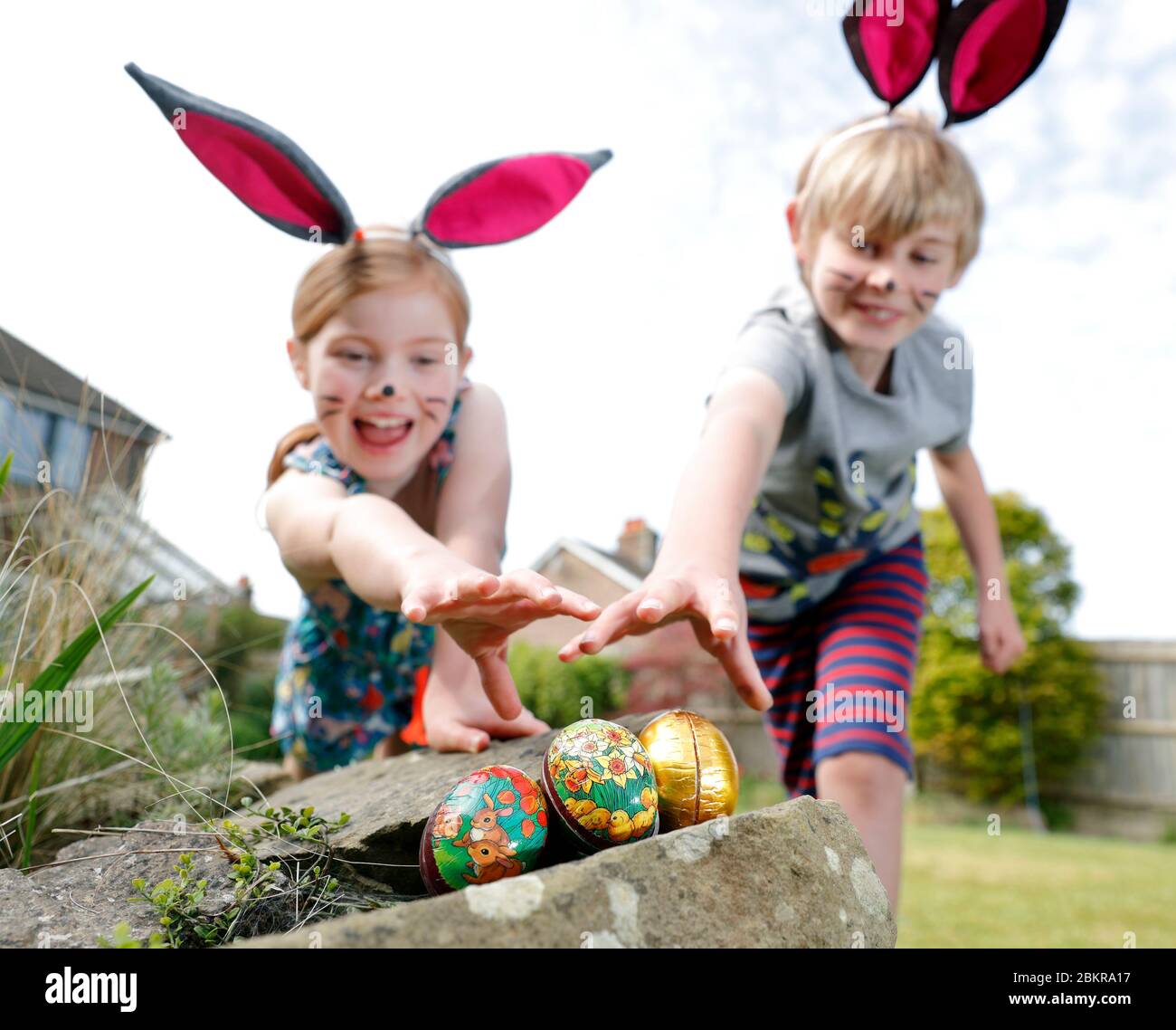 Un fratello e una sorella, indossando la pittura facciale e le orecchie conigliate fatte in casa, prendere parte a una caccia all'uovo di Pasqua la Domenica di Pasqua nel loro giardino. Foto Stock