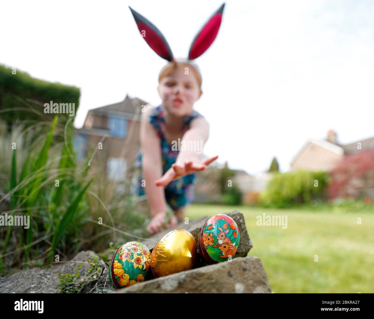 Una giovane ragazza che indossa la vernice del viso e le orecchie conigliate fatte in casa, partecipa a una caccia all'uovo di Pasqua la Domenica di Pasqua nel suo giardino. Foto Stock