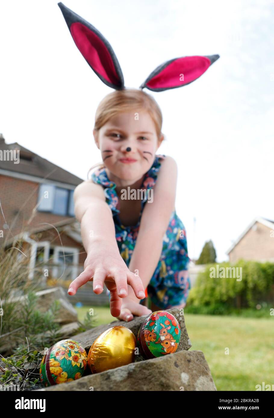 Una giovane ragazza che indossa la vernice del viso e le orecchie conigliate fatte in casa, partecipa a una caccia all'uovo di Pasqua la Domenica di Pasqua nel suo giardino. Foto Stock