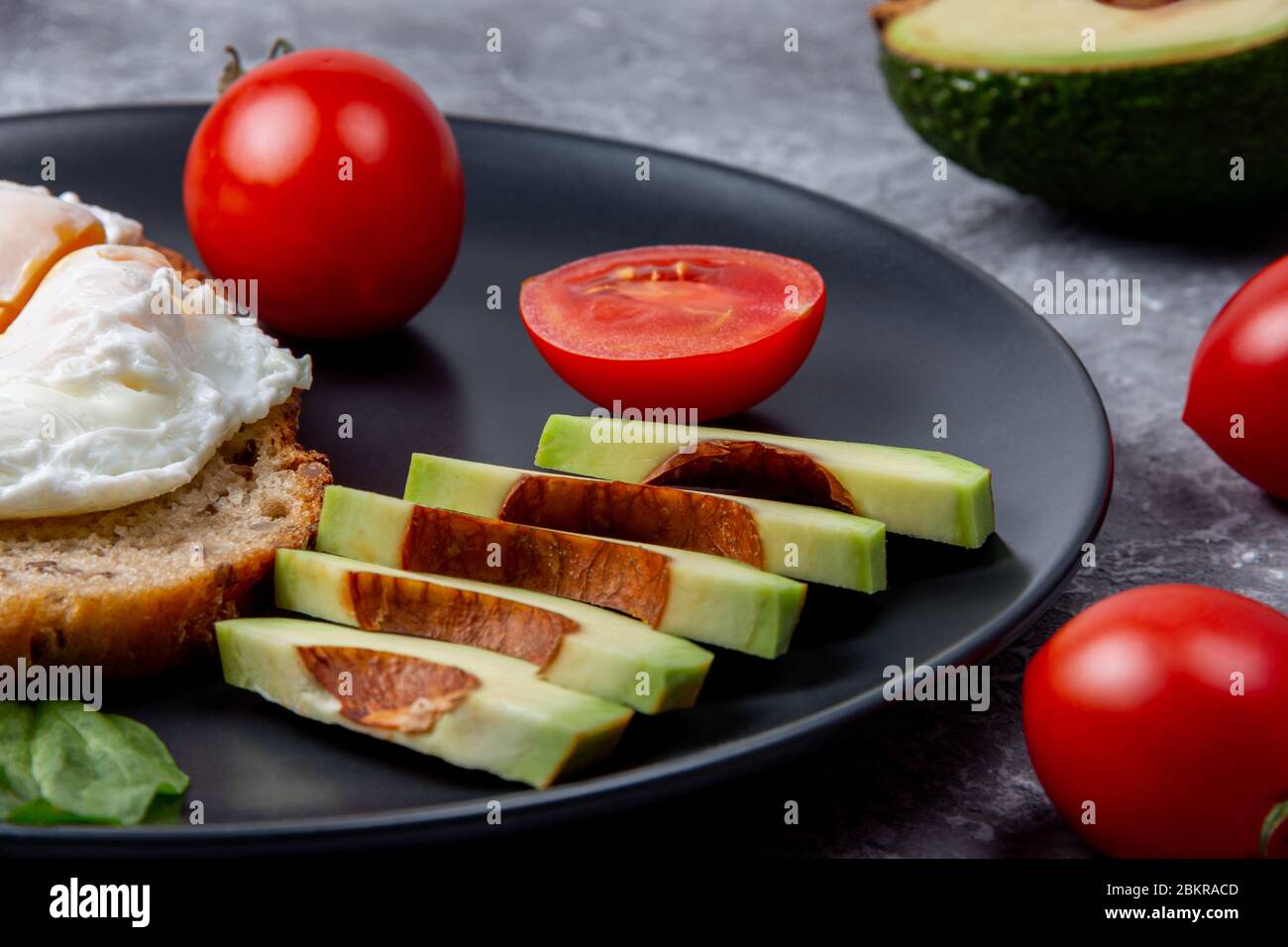 Toast avocado all'uovo in camicia con cibi salutari per la dieta su piatto nero. Pranzo ecologico con uova in camicia, pomodoro di ciliegia rossa, spinaci e avocado Foto Stock
